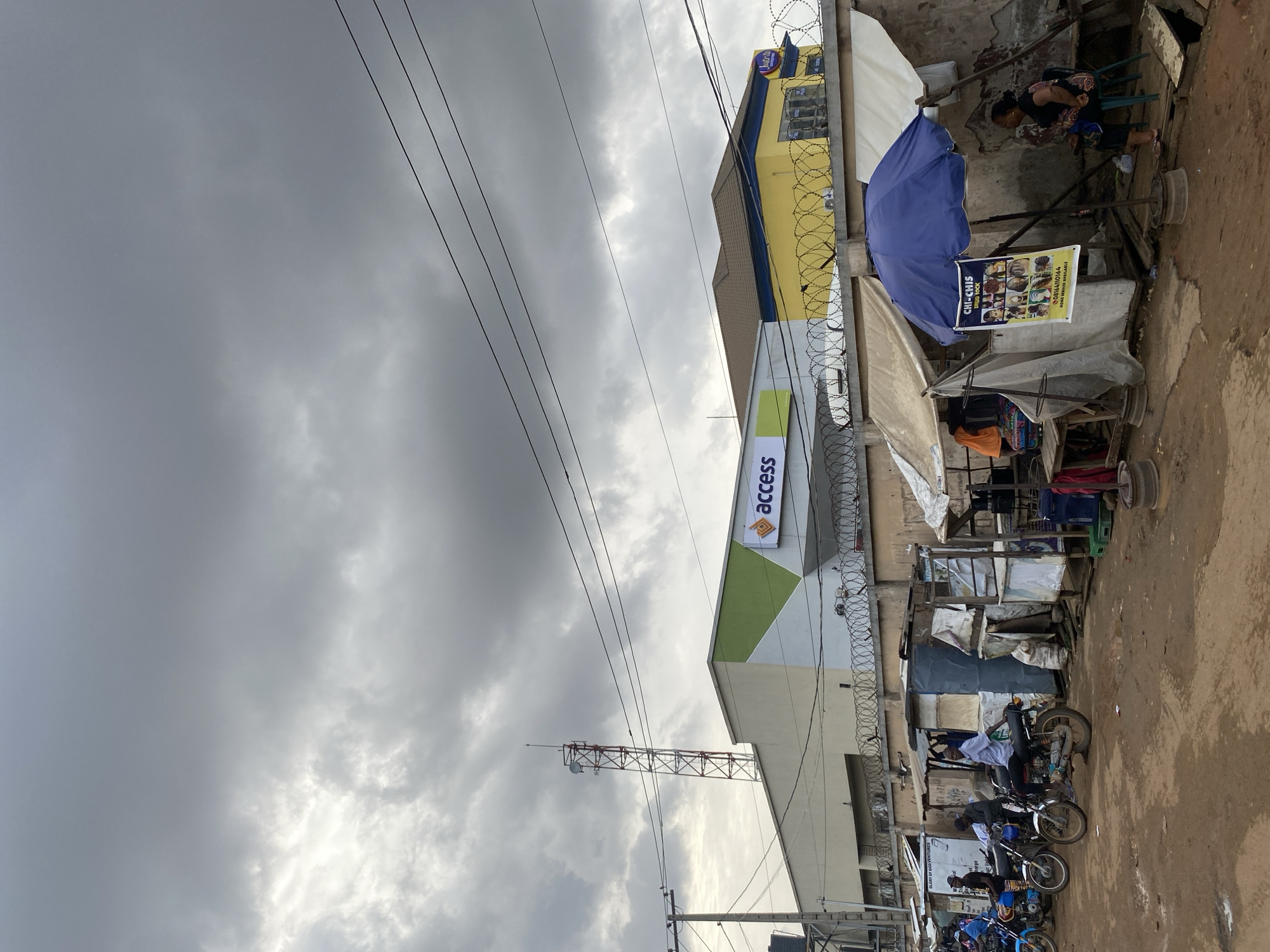 The image depicts a street scene in Lagos, Nigeria, under an overcast sky. Prominent in the frame is a building with the "access" logo, indicative of Access Bank. Adjacent to the bank, there's a yellow building with barbed wire on top. The street is lined with makeshift stalls and shops. People can be seen sitting or standing near these stalls, with a few motorcycles parked along the roadside. A communication tower is visible in the background.