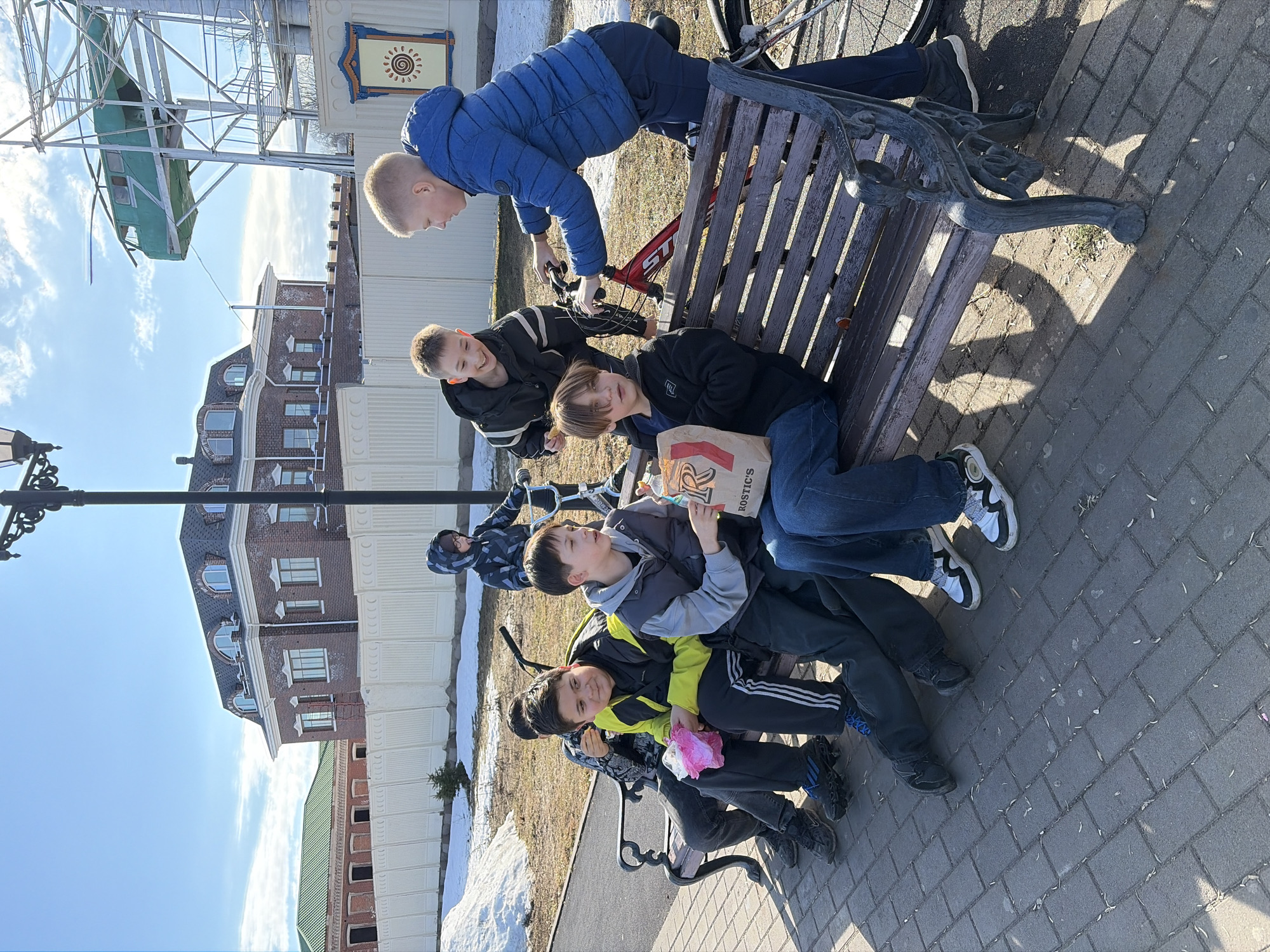The image shows a group of six young boys seated on a wooden bench in Sarapul, Russia. One boy stands behind the bench, holding a red bicycle. The boys are wearing various jackets and casual clothing suitable for cool weather. One boy holds a paper bag with "ROSTIC'S" branding. The backdrop features brick buildings with dormer windows, remnants of snow on the ground, a lamppost, and what appears to be a Ferris wheel structure. The ground is paved with brick.
