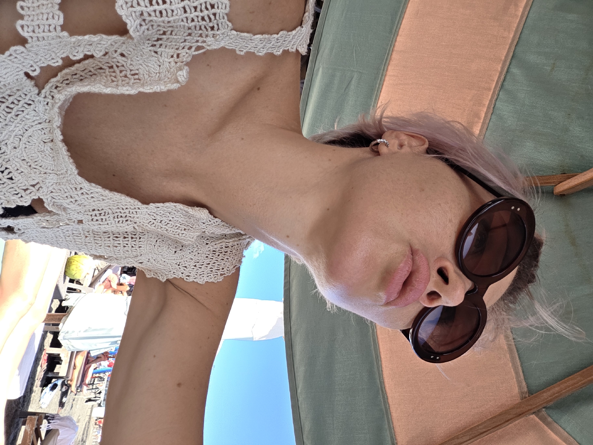 A low-angle, outdoor, waist-up shot shows a woman with light pink hair wearing large, round, dark sunglasses and a white crochet top. She is looking upwards, and her lips are pursed. She is wearing a silver hoop earring in her visible ear. The background shows a beach umbrella made of green and peachy fabric, and beyond that, a bright blue sky and a beach scene with lounge chairs and people. The location is specified as North Kuta, Indonesia.