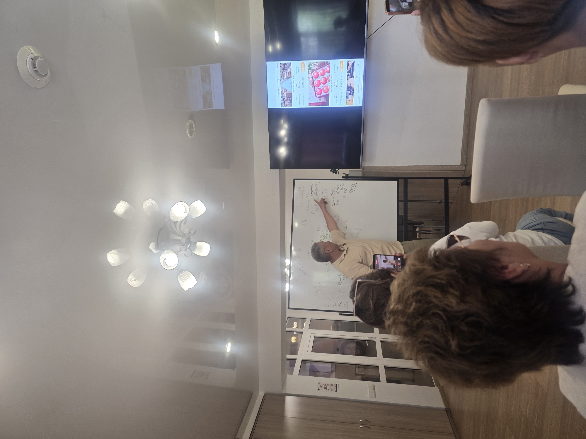 The image shows an indoor scene, possibly a conference room or classroom, with several people present. In the center of the frame, a man in a light-colored shirt is standing and pointing at a whiteboard with writing and diagrams on it. To the right of the whiteboard, a large flat-screen television displays content, which appears to be an advertisement or presentation with images of fruit. In the foreground, the backs of several people's heads are visible, indicating they are seated and facing the speaker. One person in the front is holding up a smartphone, seemingly recording the presentation. The room has a modern ceiling with recessed lighting and a chandelier. There are also wooden cabinets and glass doors visible in the background. The overall atmosphere suggests an educational or business presentation is taking place. No specific details indicating a location in Ulan-Ude, Russia are present in the image.