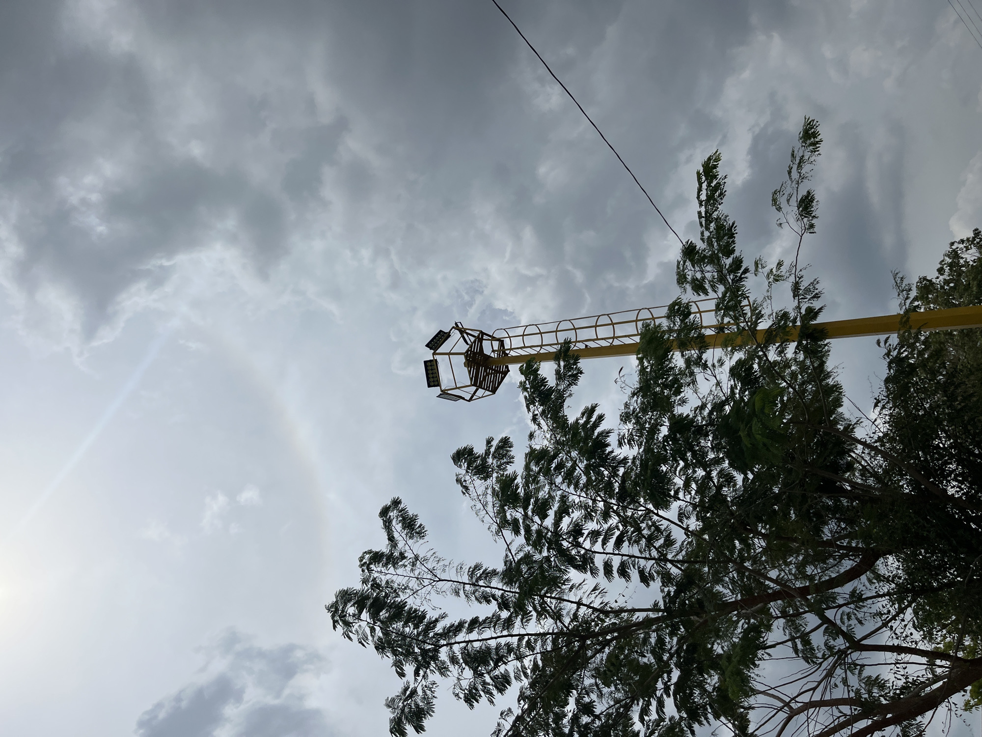 The image, taken from a low angle, shows a cloudy sky in Tinaquillo, Venezuela. A yellow, metal structure with a cage-like platform at the end is visible, possibly part of an industrial crane or lighting rig. An electrical wire crosses the sky. Trees with dense foliage occupy the lower right portion of the frame. A faint solar halo surrounds the sun in the lower left of the sky. The lighting is overcast.