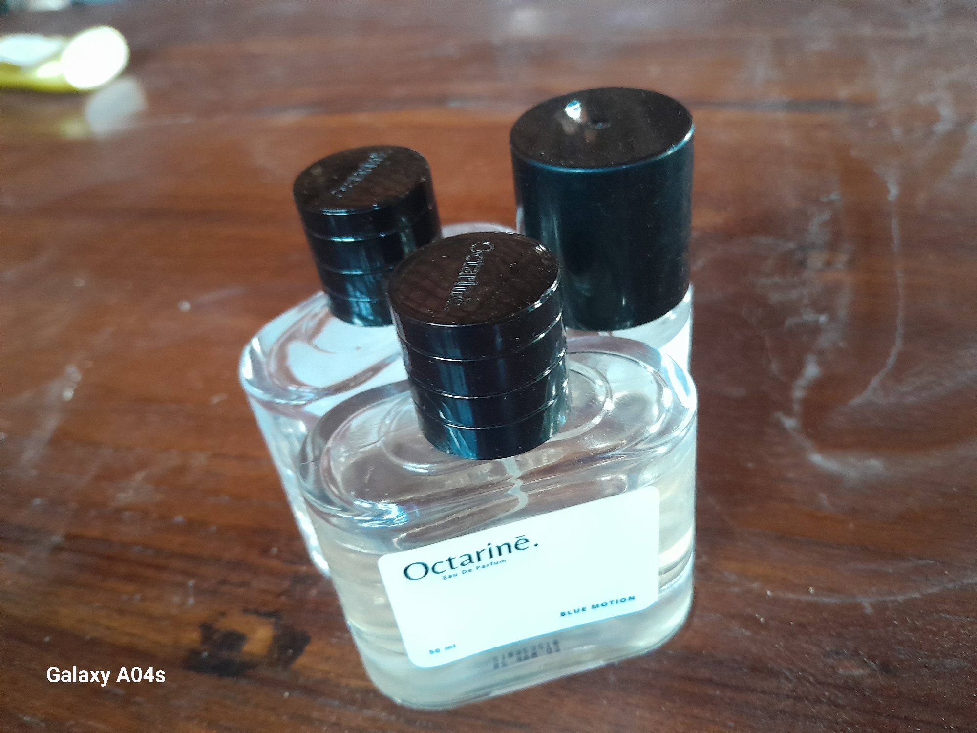 The image shows three perfume bottles on a wooden surface. The bottles are clear glass with black caps. One bottle in the foreground has a white label that reads "Octarine. Eau De Parfum BLUE MOTION 50 ml". The wooden surface appears to be a table and is dusty. The text "Galaxy A04s" is visible in the bottom left corner, indicating the device used to take the photo. There are no people or any other distinguishing environmental features visible in the image that would suggest a specific location within Pilangpayung, Indonesia.