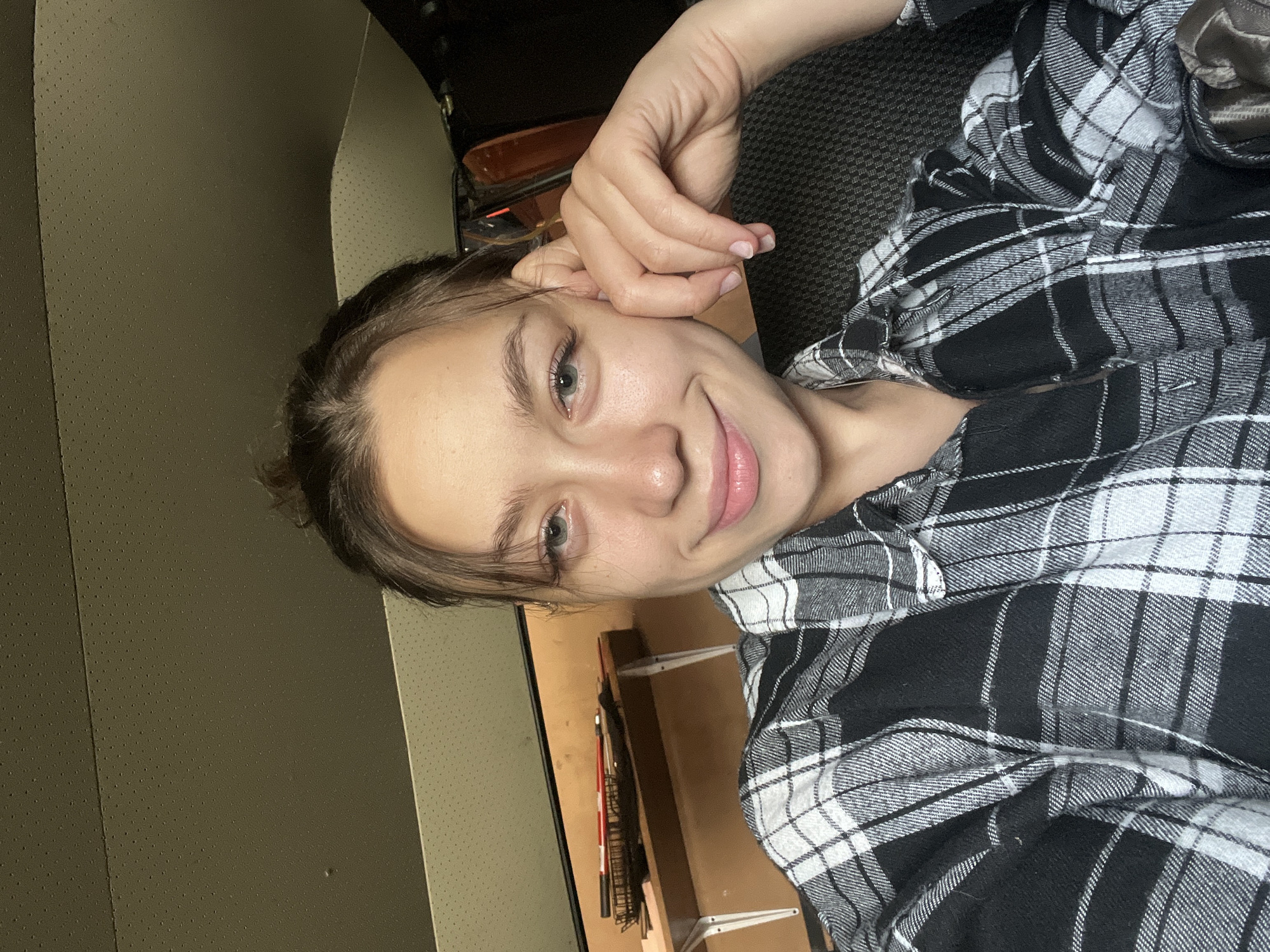 A young Caucasian woman with her hair pulled up in a bun is taking a selfie indoors, likely in a vehicle. She is wearing a black and white plaid shirt and resting her hand on her cheek, smiling at the camera. The background shows the interior of what appears to be a car, with a textured ceiling and a shelf in the background with some objects on it. The lighting suggests it is daytime, but the indoor setting makes it difficult to determine the specific location within the city of Kemul, Russia without further context.