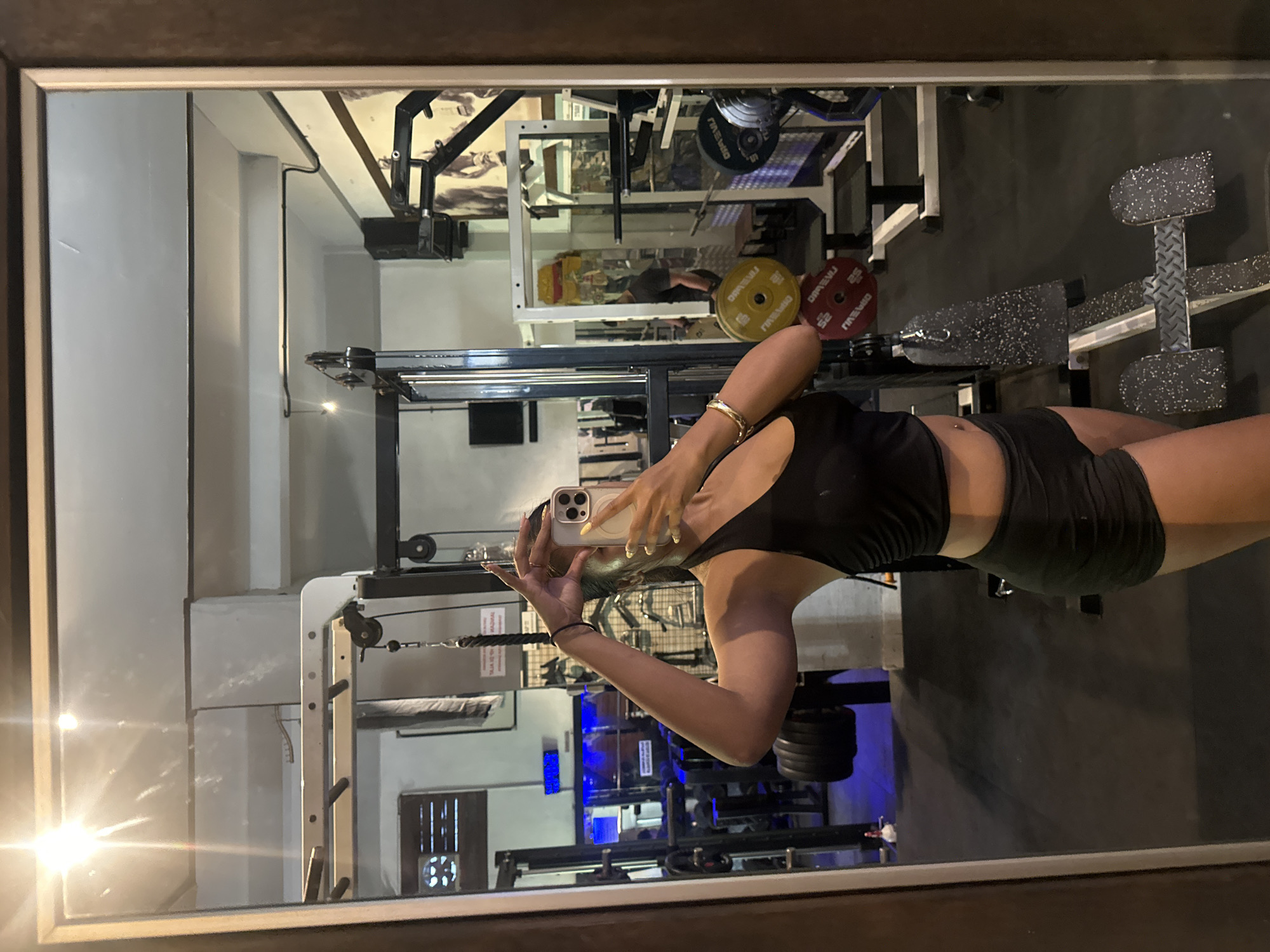 A young woman is taking a mirror selfie in a dimly lit gym in Denpasar, Indonesia. She is wearing a black sports bra and black shorts, and her hair is pulled back. She is holding her phone up to take the picture, and her other arm is bent at the elbow. In the background, gym equipment such as weight racks and benches are visible. The gym appears to be located in a somewhat older building, with exposed pipes and a textured ceiling. The overall impression is of a functional, no-frills fitness facility.