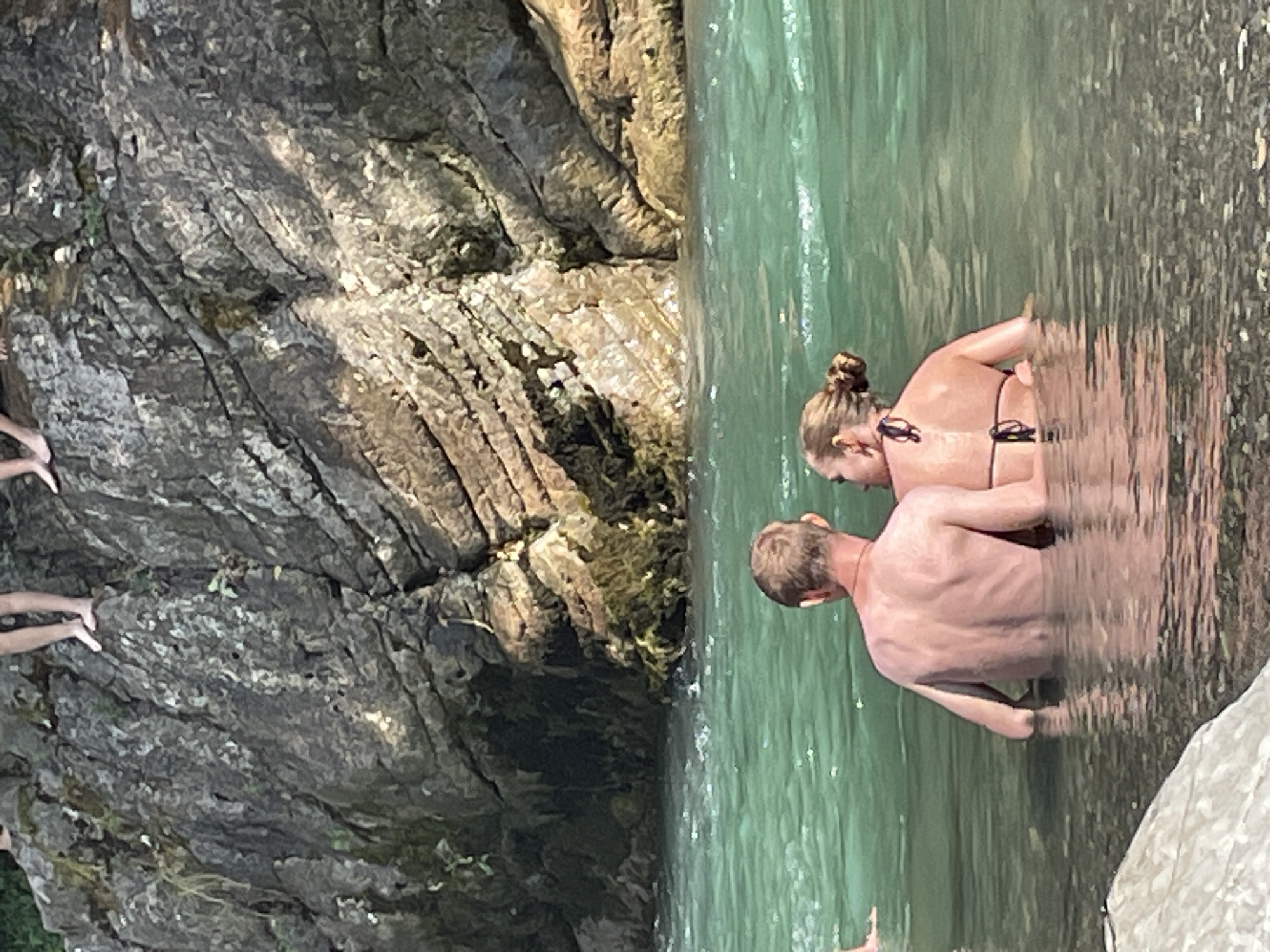 The image shows a couple in a body of turquoise water with a large, rocky cliff face in the background. The couple is facing away from the camera, embracing in the water. The man has short blonde hair and the woman has her hair in a bun. They are both wearing swimwear. The water is clear and the bottom is visible in some areas. The cliff face is steep and covered in moss in some places. There are shadows on the cliff, suggesting the sun is high in the sky. The overall impression is one of a natural, outdoor setting, likely a river or lake, with people enjoying a swim. The provided hints show cropped sections of the image, focusing on the rock formations and the people in the water, aiding in a closer examination of details.