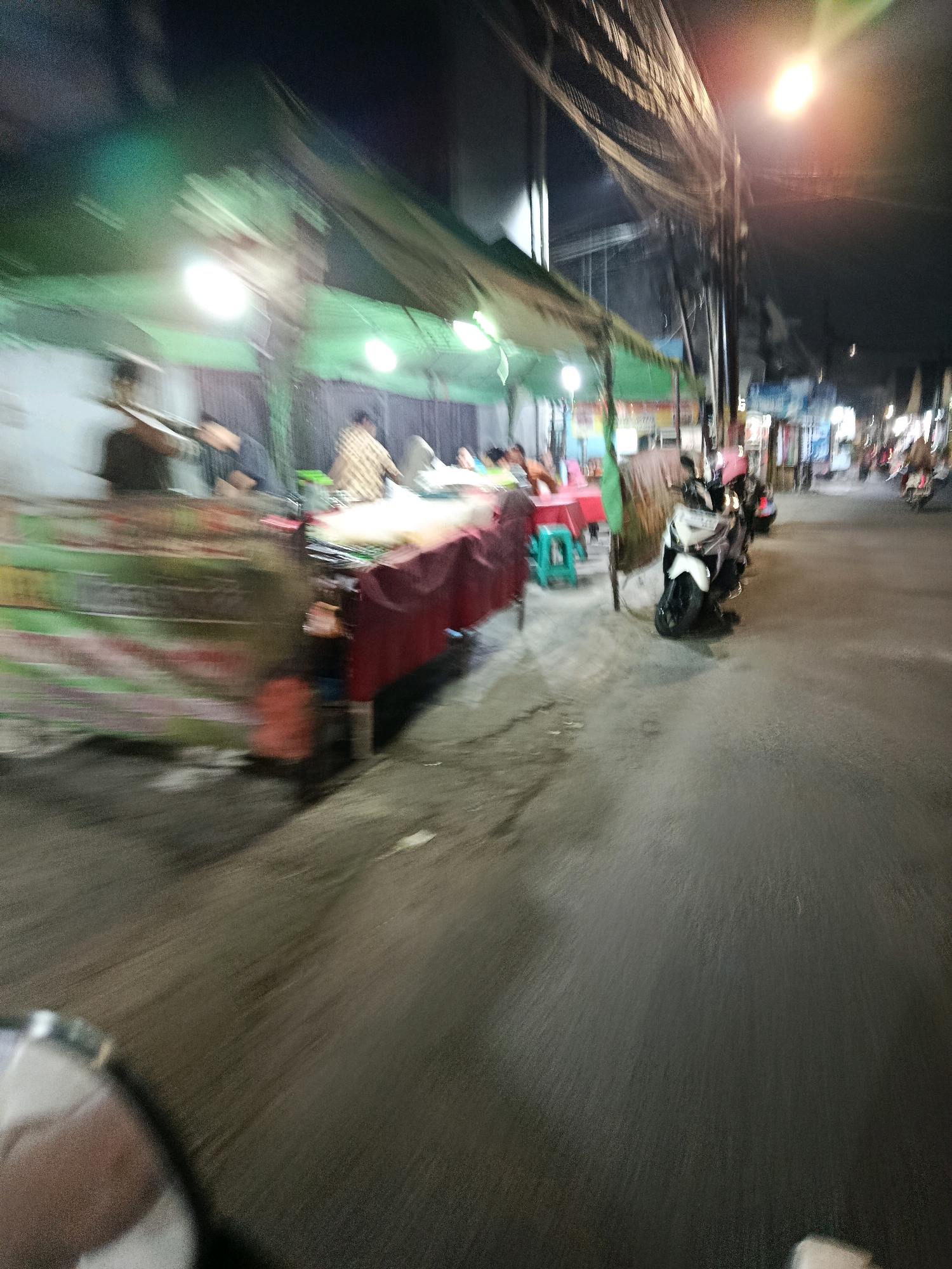 This is a motion-blurred, night-time photograph taken from a moving vehicle, likely a motorcycle, on a street in Bekasi, Indonesia. The scene depicts a row of street food stalls or small shops lining the right side of the road.
In the foreground on the left, the blurred shape of what appears to be a helmet or the upper part of a person's head is visible, indicating the viewpoint. The road surface is dark asphalt, with some patches of lighter color and faint reflections.
To the right of the road, several stalls are visible, covered by green and white awnings. Underneath the awnings, there are tables and chairs, with people seated and vendors preparing food. One stall has a red tablecloth covering a counter. Objects like bottles and bowls are present on the counters, though details are obscured by motion blur.
A white scooter is parked prominently in front of one of the stalls, also showing some motion blur. Further down the street, other scooters and possibly motorbikes are visible, along with more illuminated stalls and buildings. Overhead, a complex network of electrical wires is strung between poles, with streetlights illuminating the scene. The overall impression is of a bustling street food area at night, with the blur conveying a sense of movement and activity.