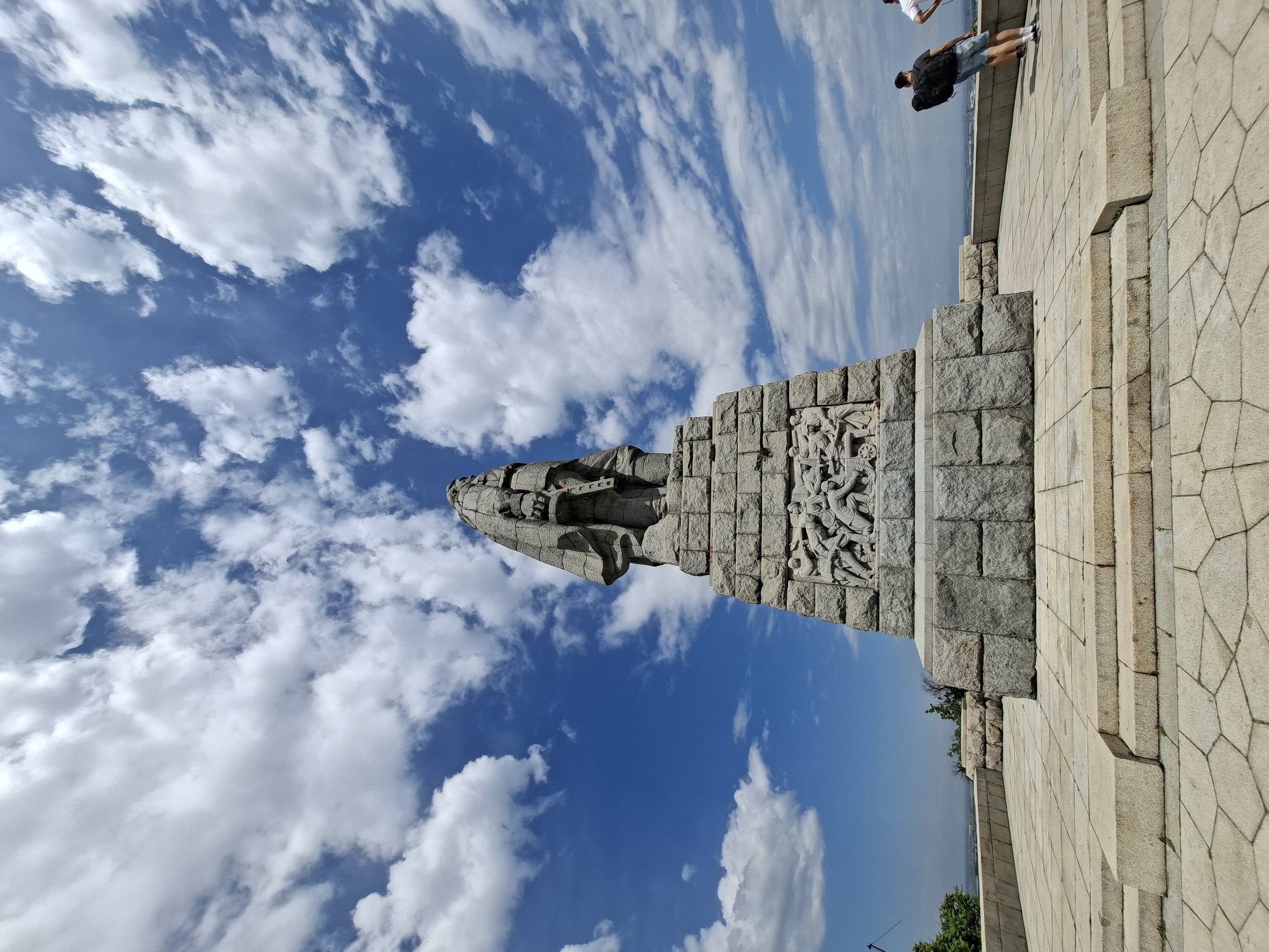 The image shows the Alyosha Monument in Plovdiv, Bulgaria. The monument is a large stone structure with a tall, stylized figure at the top, set against a background of blue sky and white clouds. Carved reliefs are visible on the side of the monument. Part of a stone plaza surrounds the monument's base. In the background, a couple of people are standing on the edge of the plaza. Vegetation is visible near the horizon.
