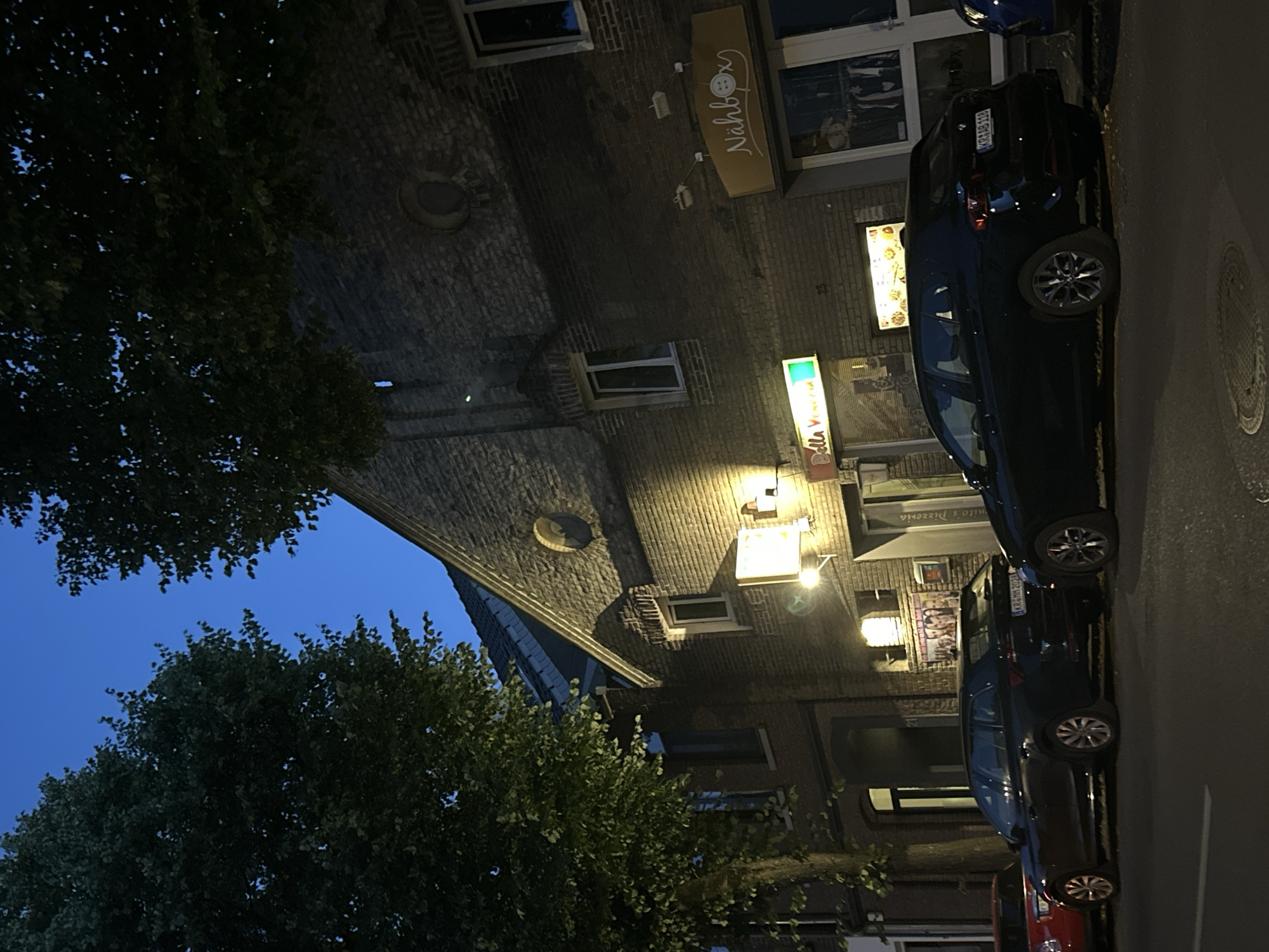 The image depicts a street scene in Krefeld, Germany, at dusk. A brick building with several windows and illuminated signs dominates the right side of the frame. One sign reads "Nähbox," and another, partially obscured, appears to be for a business named "Bella Venezia." The building's facade is dark brick, with some architectural detailing visible. Two cars are parked on the street in front of the building: a dark-colored station wagon and a red car partially visible on the left. The station wagon has a license plate that reads "KR-AB 118." Trees with dark green foliage frame the top and left portions of the image, with the sky appearing a deep blue, indicating late evening. The street is asphalt, and a manhole cover is visible on the lower right. The overall atmosphere is calm and quiet.