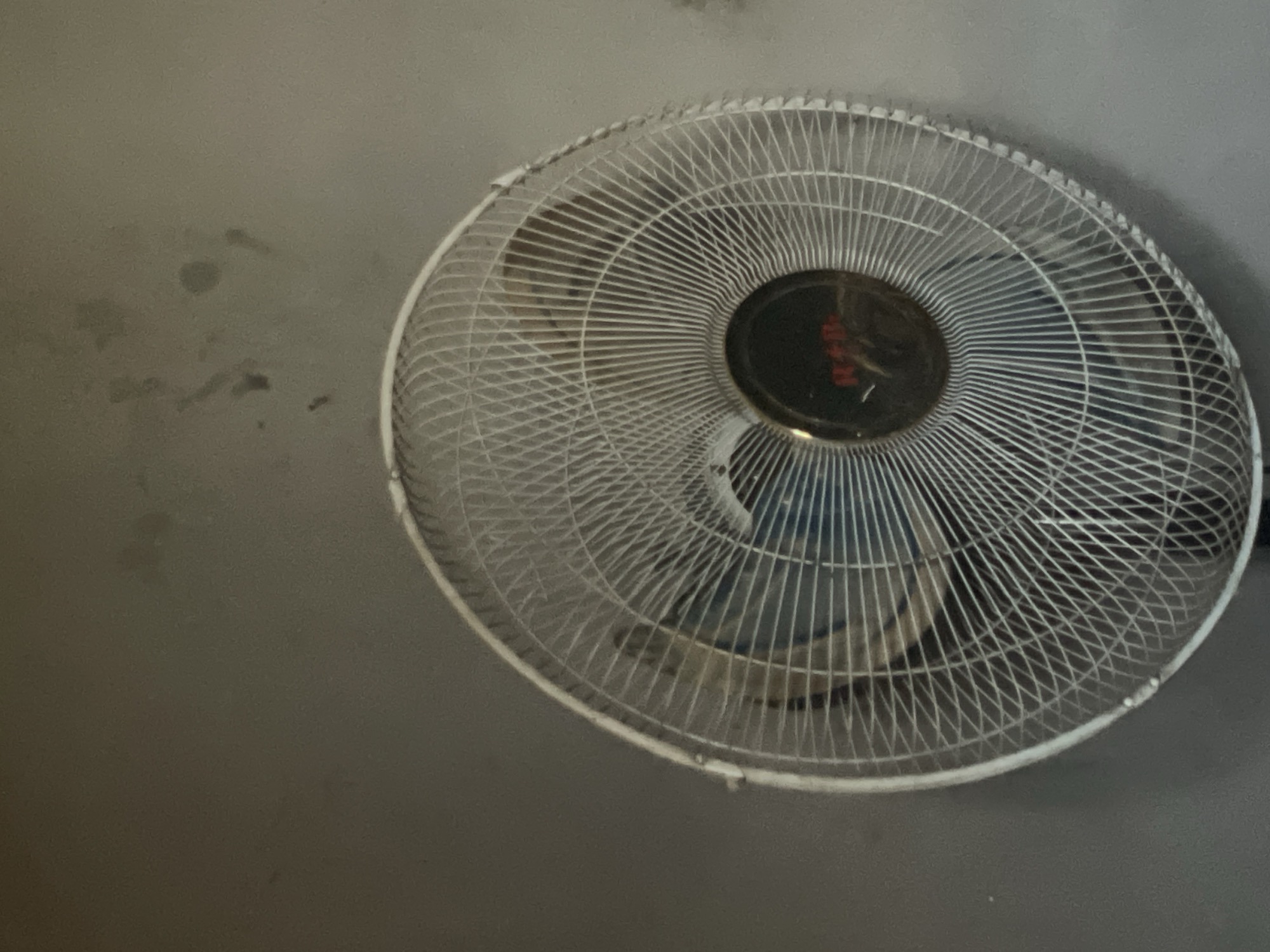 The image shows a white, circular electric fan mounted on a gray wall. The fan has a metal cage surrounding the blades, with a dark circular hub in the center. The wall has some smudges or marks on the left side. The location is unknown.