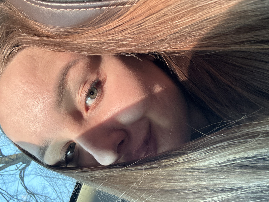 An upward-angled, close-up photograph depicts a female individual with light brown hair and green eyes, observing the camera with a subtle smile. Direct sunlight from the upper left casts a distinct shadow across the bridge of her nose and right cheek. Her long, straight hair, illuminated by the sun, frames her face. Behind her, through a window on the left, bare tree branches are visible against a blue sky. A dark brown, textured car seat is partially visible on the right. The scene is situated in Chaikovsky, Russia.
