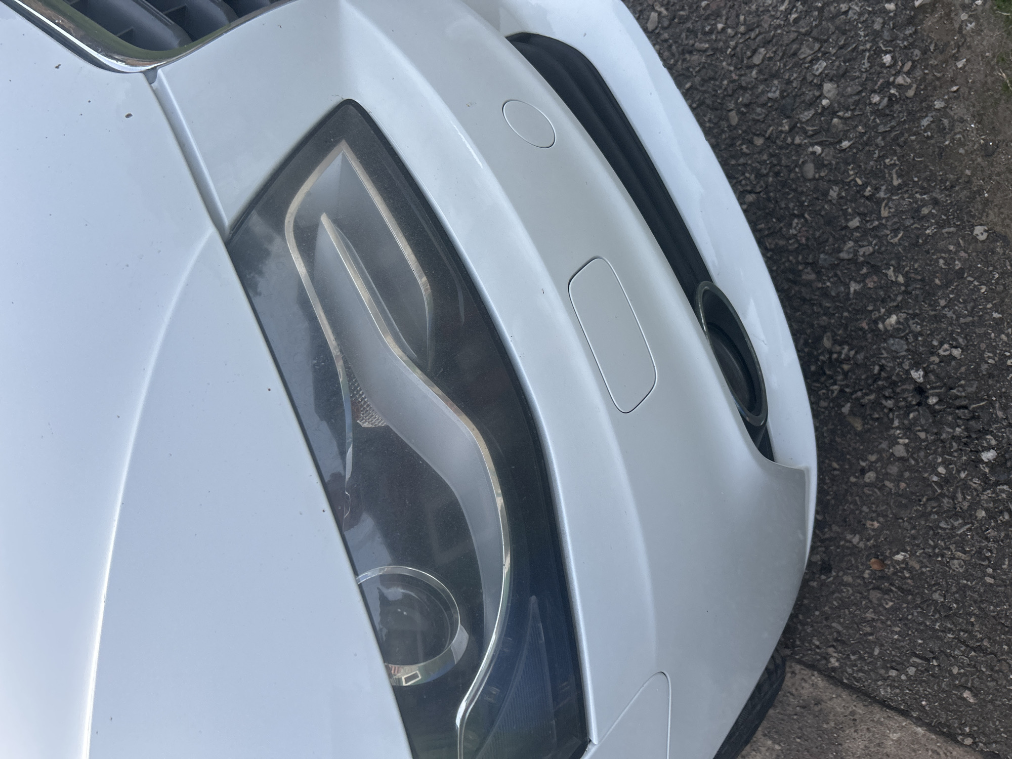 The image shows a close-up, angled view of the front right side of a white car, likely parked on asphalt. The car appears to be modern, with a distinctive headlight design featuring chrome accents and dark interior components. Part of the grille, with chrome trim, is visible at the top left. The bumper has a subtle curvature and includes a rectangular panel, possibly for a license plate mount or tow hook cover, and a circular sensor or marker. The texture of the asphalt, with visible pebbles and some dirt, suggests an outdoor setting. There are no people visible in the image. The lighting suggests it is daytime, but the weather conditions are not discernible. No text is visible in the image. The scene is likely a street or driveway in Daugavpils, Latvia, given the provided location context.