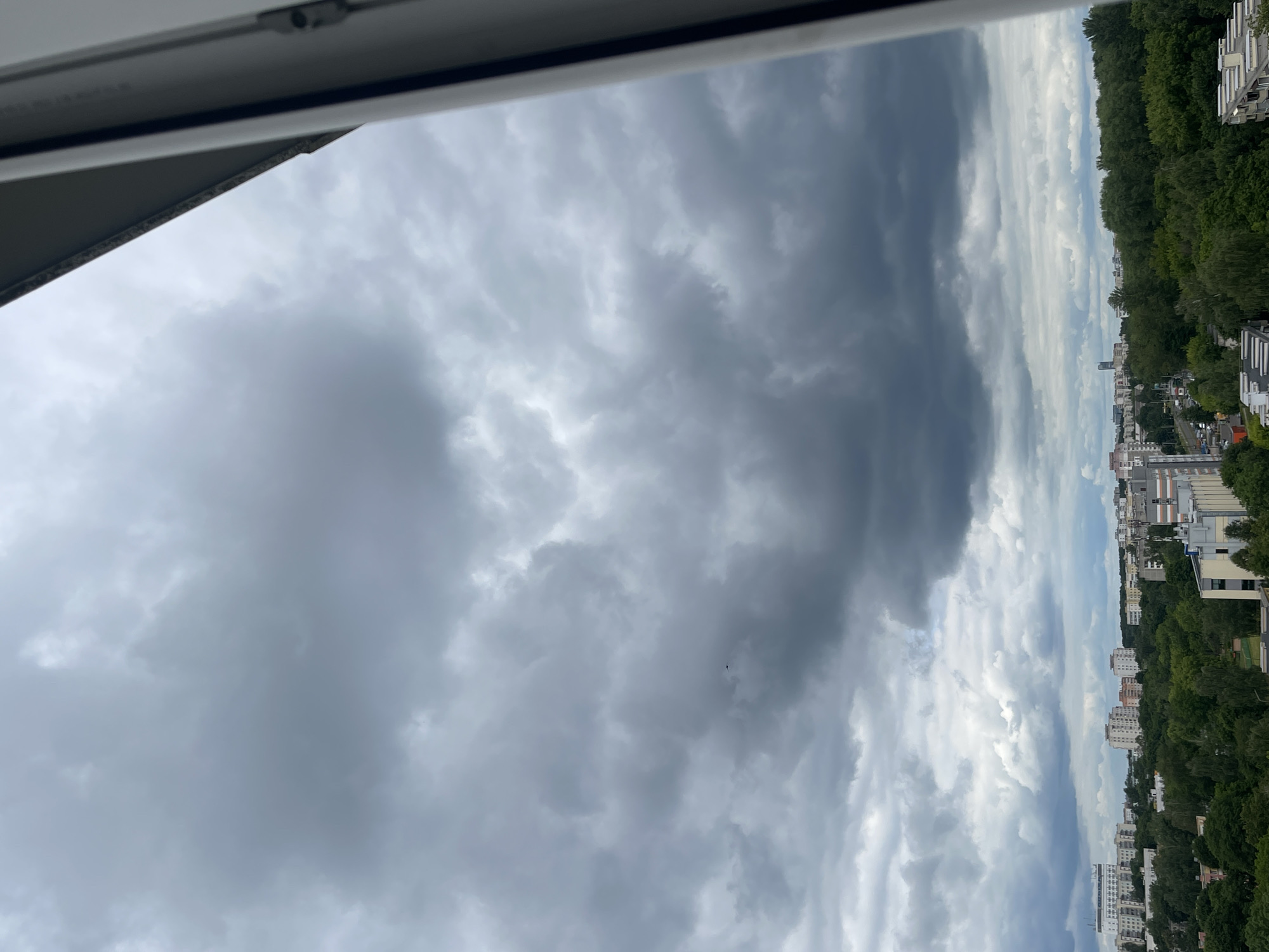 The image shows a city skyline viewed from above. The sky is filled with dark, dramatic clouds, suggesting an impending storm or heavy overcast. The cityscape below is partially obscured by lush green trees that line the horizon, with buildings of various heights interspersed among them. The architecture appears to be modern, with apartment buildings visible. The overall scene conveys a sense of atmospheric tension due to the stormy sky, contrasting with the otherwise serene depiction of urban life and nature. The time of day is difficult to ascertain precisely, but the lighting suggests it could be late afternoon or early evening. No people or specific objects are distinctly visible, with the focus being on the sky and the urban landscape. The location is identified as Minsk, Belarus.