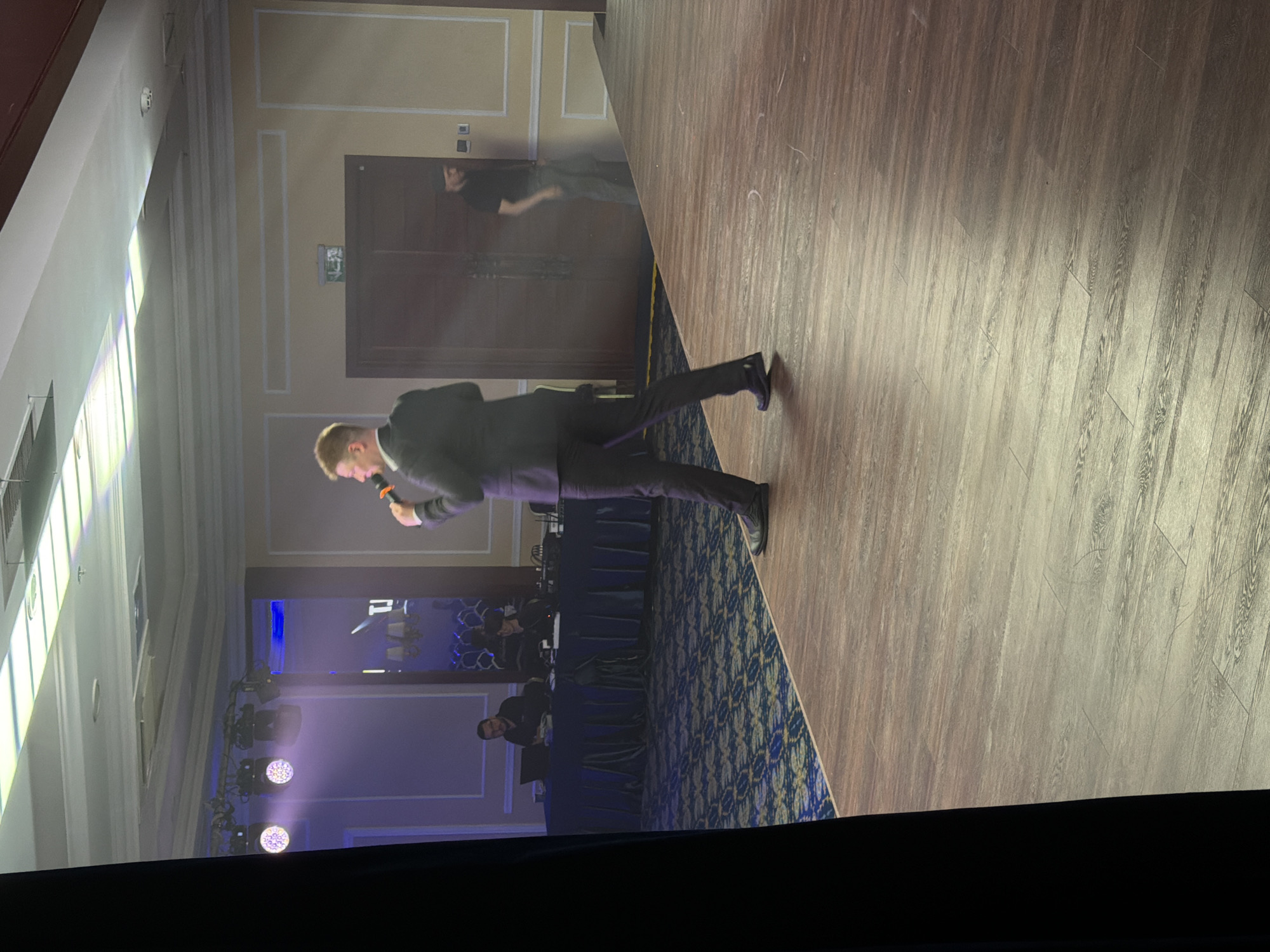 A man in a suit is speaking into a microphone while standing on a wooden stage. He is facing away from the camera. In the background, there are several people sitting at tables, and a man in a black t-shirt and pants is walking towards a door. The room is lit by stage lights. The stage is made of wood and the floor has a patterned carpet.
