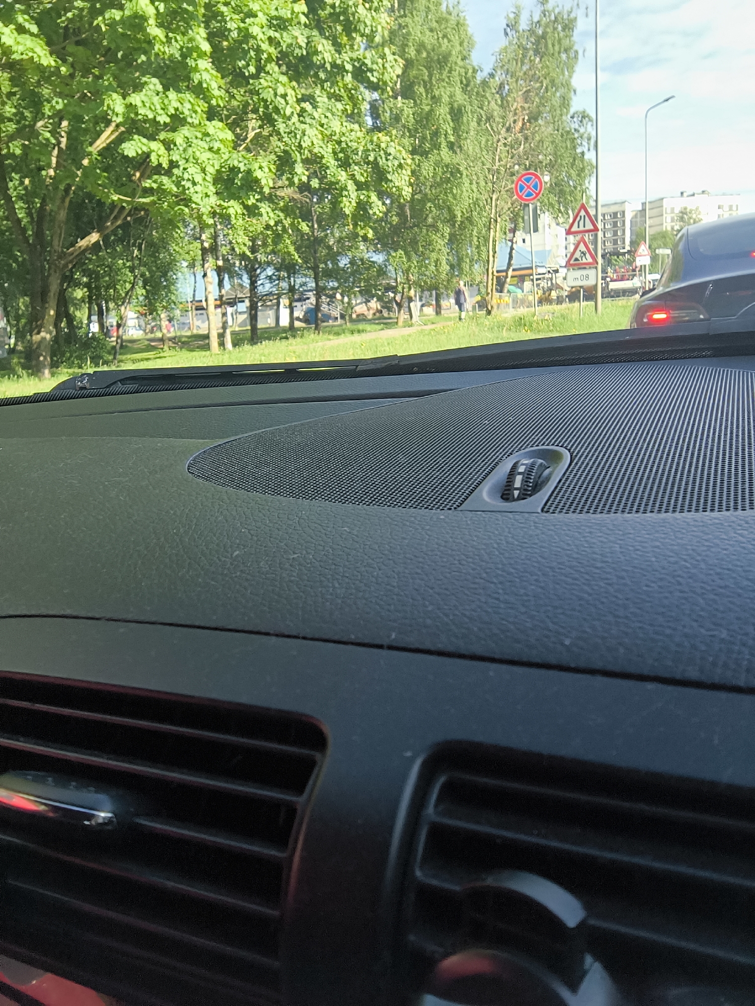 The image is taken from inside a vehicle, focusing on the dashboard and the view through the windshield. The dashboard is black, with a textured surface and a speaker grill featuring a central adjustment knob. The vehicle is stopped in traffic, behind another car whose taillight is visible. Outside, the environment includes green trees, grass, and a pedestrian walkway. In the distance, a "No Stopping" traffic sign and road work signs are visible, along with buildings. Based on these signs, the location is Riga, Latvia. There are also pedestrians on the sidewalk near construction.