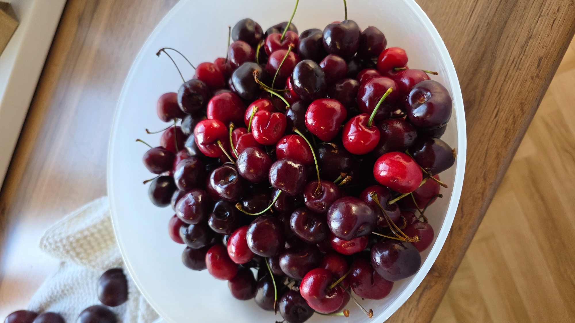 A close-up, high-angle shot shows a white bowl overflowing with ripe cherries. The cherries are a mix of deep red and dark burgundy, with some exhibiting a brighter red hue. Many of the cherries still have their green stems attached. The bowl is placed on a wooden surface, likely a table, which has a visible grain pattern. To the upper left of the bowl, a white textured cloth is partially visible, with a few dark, round objects that appear to be more cherries scattered on it. The lighting suggests an indoor environment, casting subtle shadows on the wooden surface. There are no people or animals present in the image. The scene is static, depicting a collection of fruit ready for consumption or preparation. The specific location in the city of Алга, Russia, cannot be determined from the visual information provided in the image.