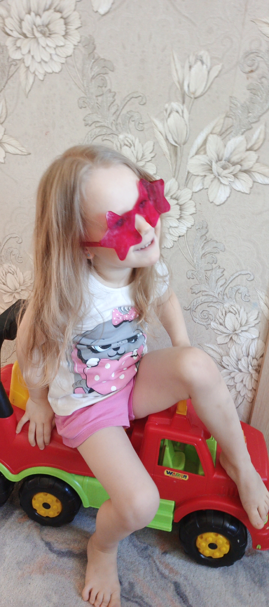 A young girl with long blonde hair is sitting on a red and green toy truck, positioned indoors against a textured wall with a floral pattern. She is wearing pink star-shaped sunglasses, a white t-shirt with a cartoon cat graphic, and pink shorts. The girl is smiling and looking to her right. Her bare feet are visible, with one foot resting on the red hood of the toy truck. The background wallpaper has raised floral designs in neutral colors. The location is Chaikovsky, Russia.