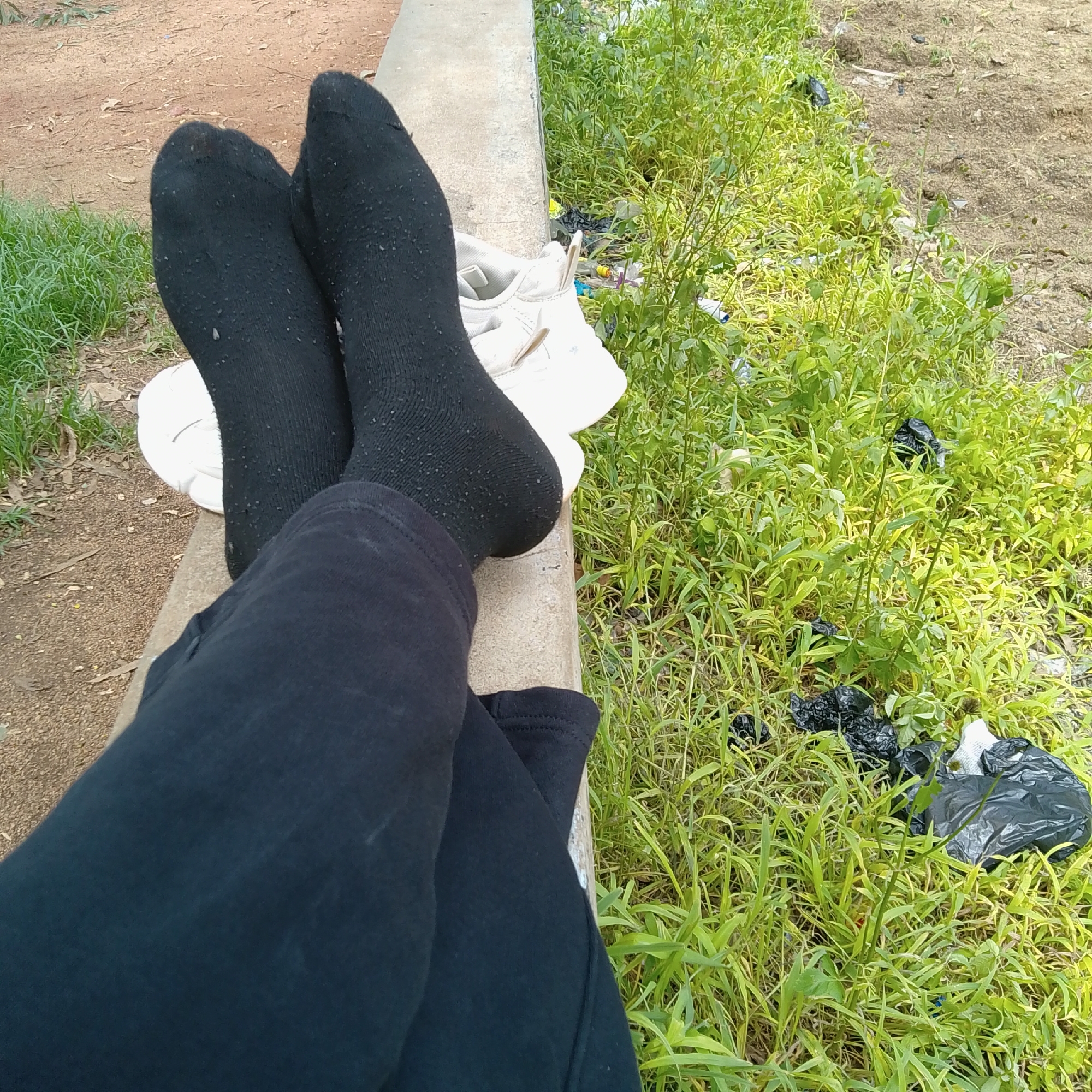 The image shows a close-up view of a person's legs and feet, which are clad in black socks and dark pants. The person is resting their feet on a concrete ledge, with a white sneaker partially visible underneath. The background is a mix of dirt, sparse green grass, and some scattered black plastic bags, suggesting an outdoor, potentially urban or semi-urban environment. The lighting indicates it is daytime.