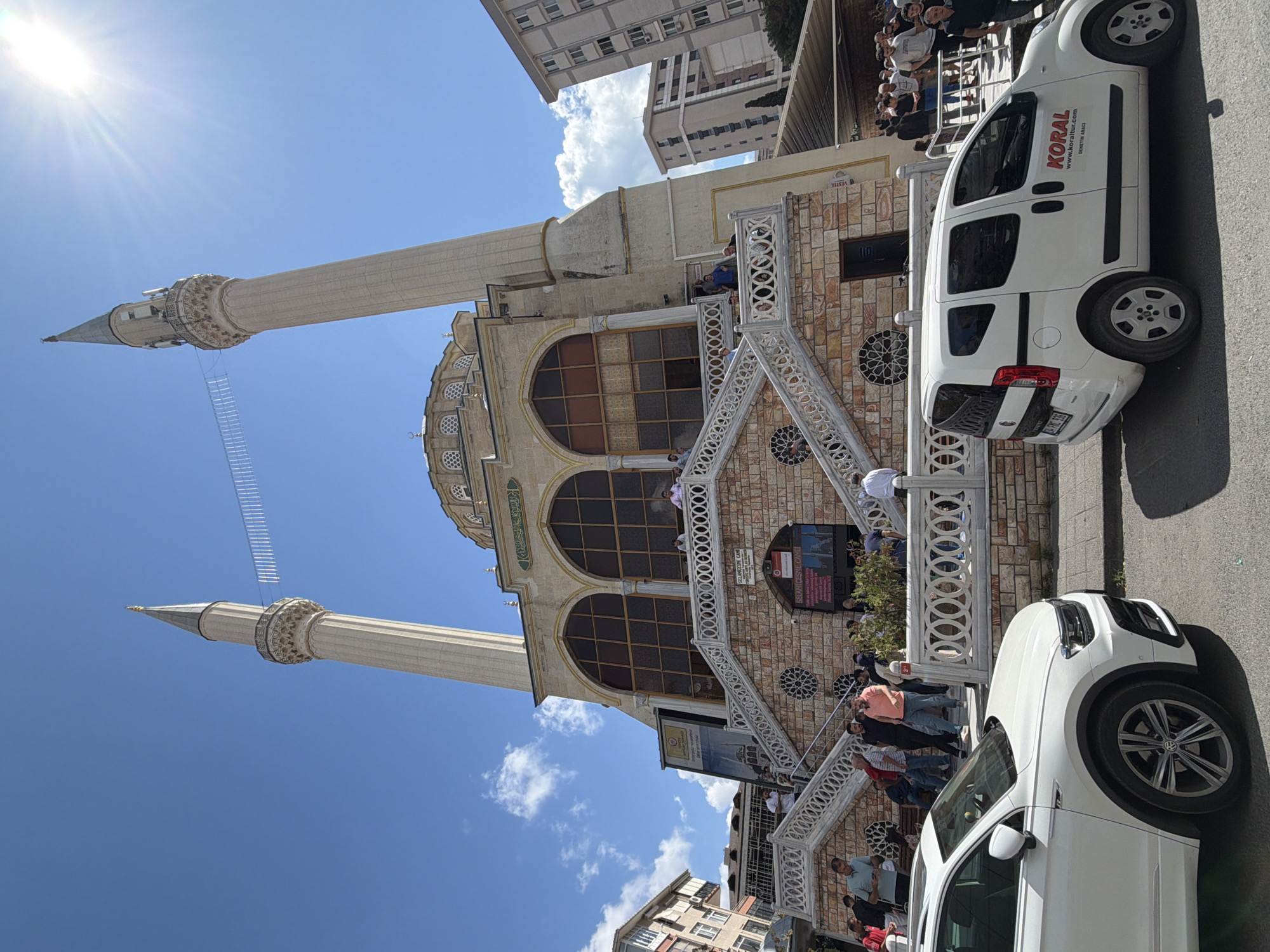 The image shows a mosque with two minarets in Kadıköy, Turkey. The mosque is made of stone and has a dome. There are people gathered in front of the mosque, some are walking up the stairs, while others are standing around. Two white vehicles are parked in front of the mosque. The sky is blue and there are some clouds. The sun is shining brightly, casting shadows on the ground. A banner with Turkish text is displayed on the mosque's exterior. There are buildings in the background.