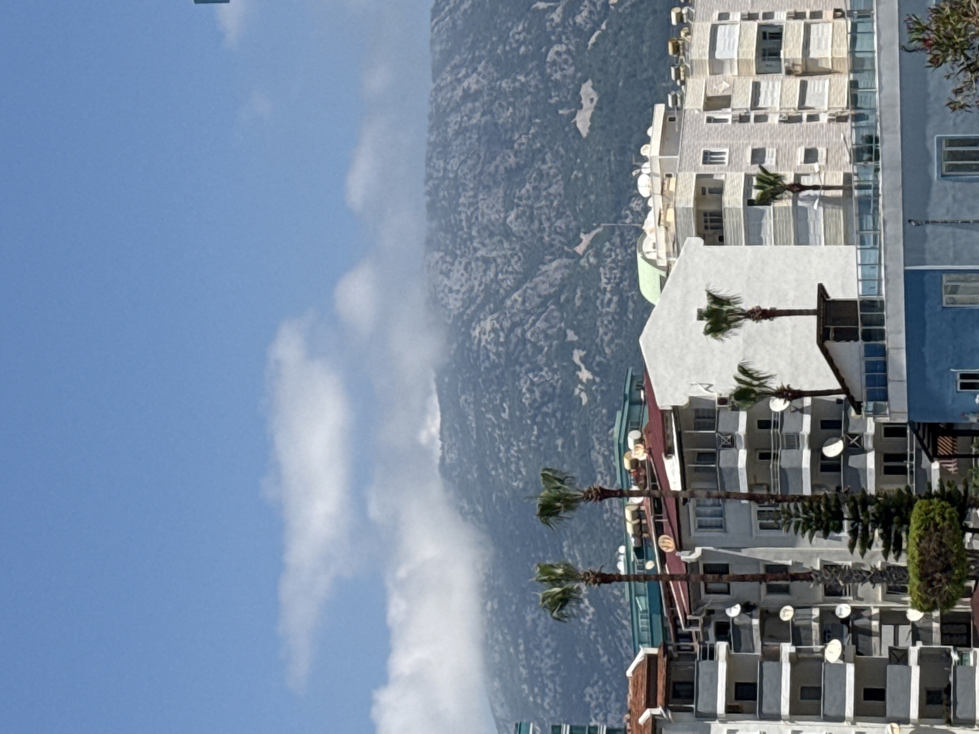 The image captures a vibrant daytime scene featuring urban structures set against a mountainous backdrop. The perspective is slightly elevated, looking towards a sprawling landscape.
In the foreground and midground, a cluster of multi-story buildings dominates the right side of the frame. These structures appear to be residential or hotel complexes, characterized by numerous windows, balconies, and a prominent display of individual satellite dishes affixed to their exteriors and rooftops. One building is predominantly white, another a light grey, and a third on the far right is a distinct blue. Several tall palm trees with green fronds are scattered amongst these buildings, adding a tropical touch to the environment. Other green vegetation, possibly bushes or smaller trees, is also visible near the buildings.
Stretching across the center and left of the image is a large, rugged mountain range. The mountains have a rocky, textured surface, appearing dark grey under the sunlight. A thin layer of white clouds or haze subtly obscures the lower slopes of these mountains, creating a soft transition between the sky and the terrain.
The sky, occupying the upper left portion of the image and above the mountains, is a clear, bright blue with scattered, wispy white clouds. The overall lighting suggests a sunny day, likely morning or afternoon, indicating fair weather conditions. There is no discernible human activity or visible text in the scene. The combination of modern buildings, palm trees, and prominent mountains suggests a location in a coastal or resort area.
