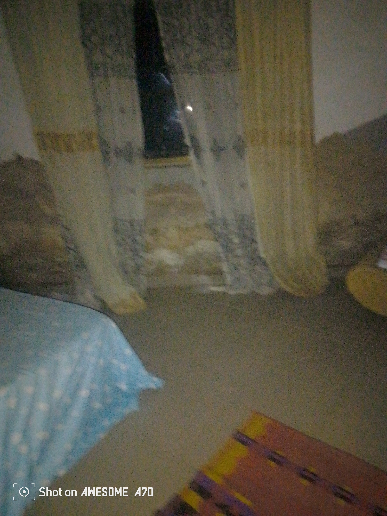 The image shows a dimly lit bedroom with a bed in the foreground partially covered by a light blue blanket with white star-like patterns. Curtains hang on the right side of the frame, appearing to be made of sheer fabric with a decorative pattern. Behind the curtains, a window is visible, revealing a dark exterior with a faint light source. The floor is covered with a carpet. On the lower right side of the image, a portion of a rug with yellow and purple geometric patterns is visible. A watermark on the bottom left indicates the photo was "Shot on AWESOME A70".