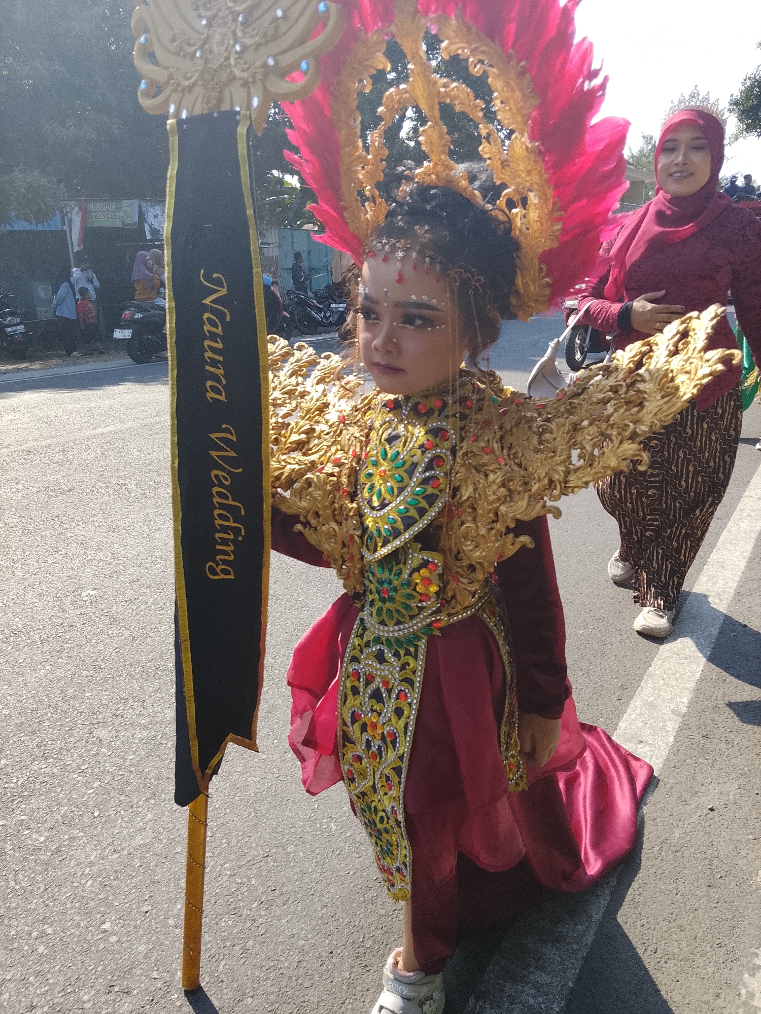 In Buduan, Indonesia, a young girl in a vibrant, ornate costume, possibly for a parade or celebration, is the central focus of the image. Her outfit features a gold headdress adorned with pink feathers, elaborate gold shoulder pieces, and a richly decorated sash worn diagonally across her chest. She is holding a tall black banner with the words "Naura Wedding" written in gold script. Beside her, a woman wearing a maroon hijab and a patterned maroon dress walks with her hand on her hip, smiling. The scene takes place on a paved road, with a white line marking the edge of a pedestrian walkway or traffic lane. In the background, trees and buildings are visible, suggesting an outdoor urban or semi-urban setting. Several motorcycles are parked or being ridden in the distance, and other people are present, some observing the procession. The bright sunlight casts distinct shadows on the road, indicating it is daytime.