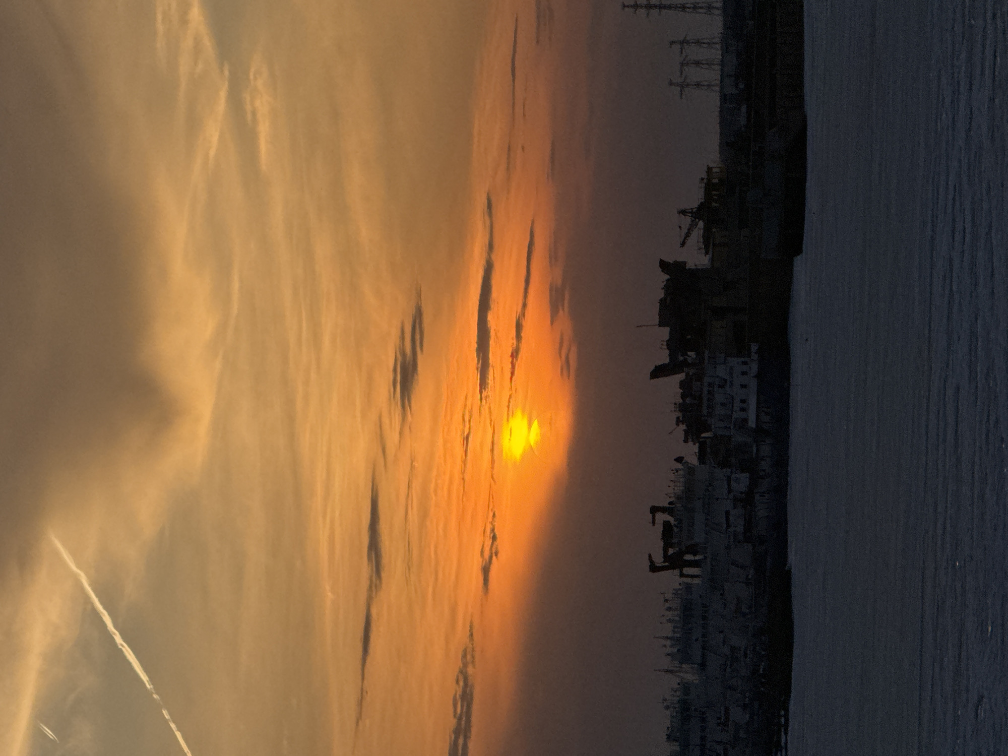 The image shows a sunset over what appears to be a body of water, likely a reservoir, in the city of Chaikovsky, Russia. The sun is positioned on the right, casting a bright orange glow through the clouds. Silhouetted against the sunset is the outline of the city's industrial area, featuring buildings, cranes, and power lines. The sky varies in color from pale yellow to deep orange, with dark clouds providing contrast. The water appears dark and still, reflecting the colors of the sunset. The image is oriented sideways.
