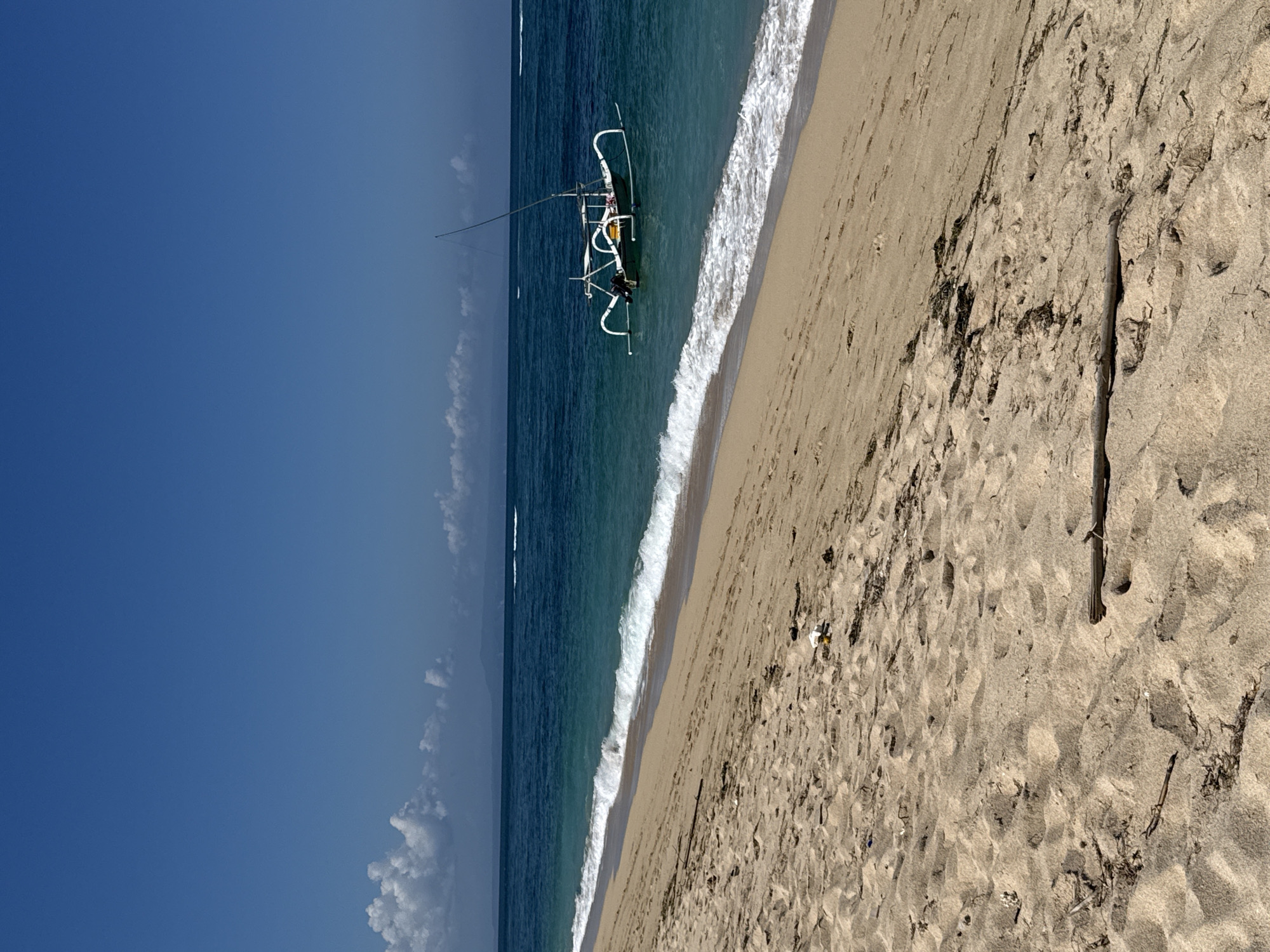 A wide shot captures a serene beach scene in Kuta Selatan, Indonesia. The foreground is dominated by textured, light-brown sand, scattered with dried seaweed and a piece of driftwood. The middle ground features the gentle turquoise waves of the ocean lapping at the shore, creating a white foamy line. Further out, a traditional Balinese boat with outriggers, painted white, floats calmly on the clear blue water. In the background, a distant, hazy coastline is visible under a clear, bright blue sky, with a few wispy white clouds scattered towards the horizon. The overall atmosphere is tranquil and sunny, conveying a sense of peaceful seaside solitude.