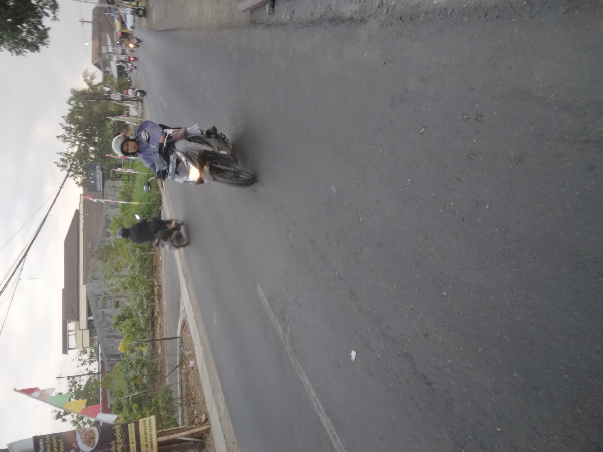 In the city of Desa Pamijen, Kecamatan Sokaraja, Indonesia, a person on a motorcycle is seen riding on an asphalt road. The person is wearing a helmet and a blue rain poncho. The motorcycle is dark colored with its headlight on. In the background, there are several other motorcycles, buildings, trees, and flags. The sky is overcast.