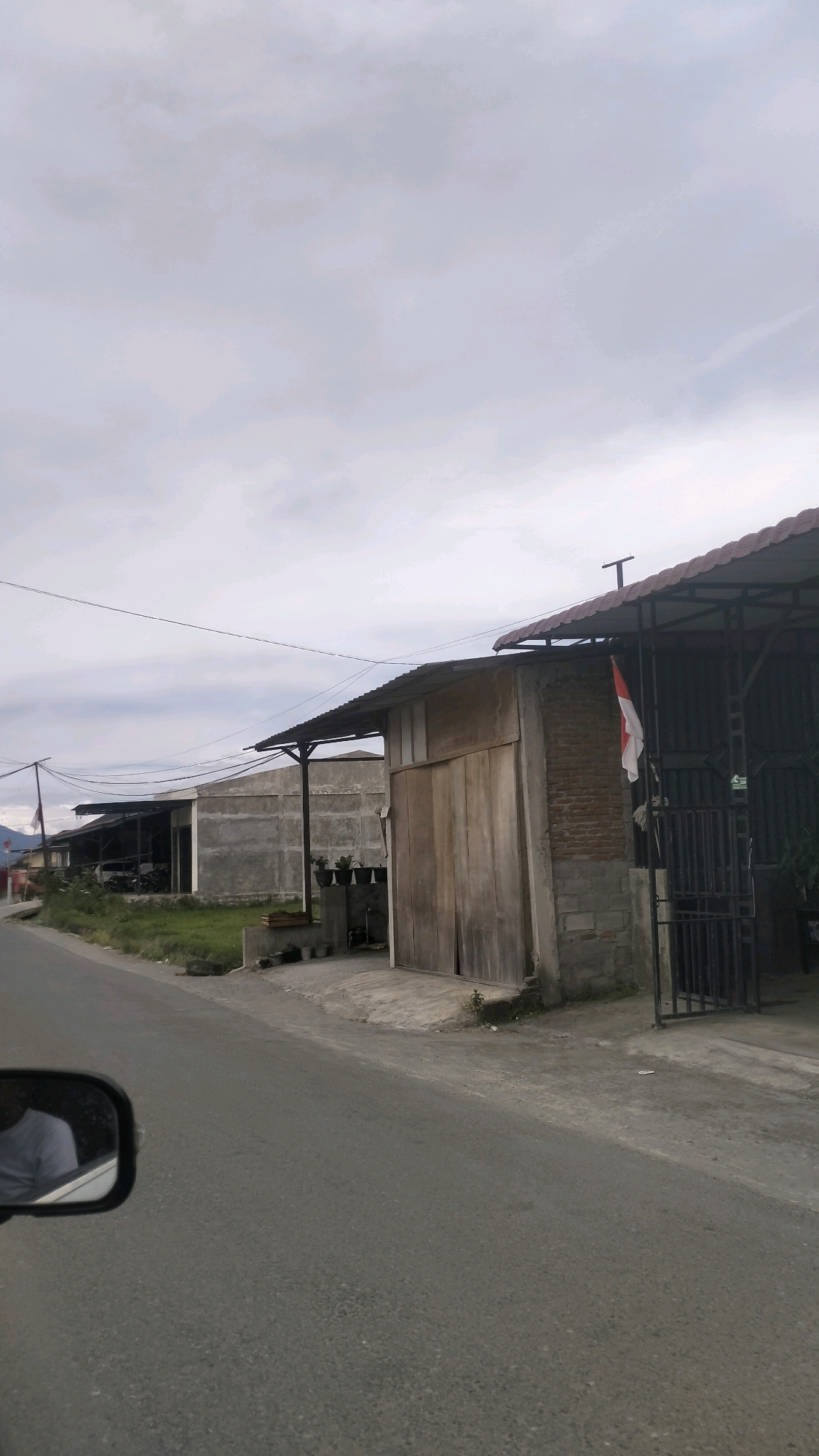 The image shows a street scene in Lotkala, Indonesia. The foreground features a grey asphalt road with a car's side mirror on the left, reflecting a person wearing a light blue shirt. Beyond the road, there are several buildings with open-sided roofs, typical of structures in this region. One building on the right has a prominent wooden door and a brick section, with an Indonesian flag hanging vertically. Several potted plants are visible near this building. In the background, more concrete buildings with corrugated metal roofs line the street, and there is a grassy area between the road and the structures. The sky is overcast with grey clouds. The scene appears to be a commercial or industrial area, possibly a workshop or storage facility, given the nature of the buildings.