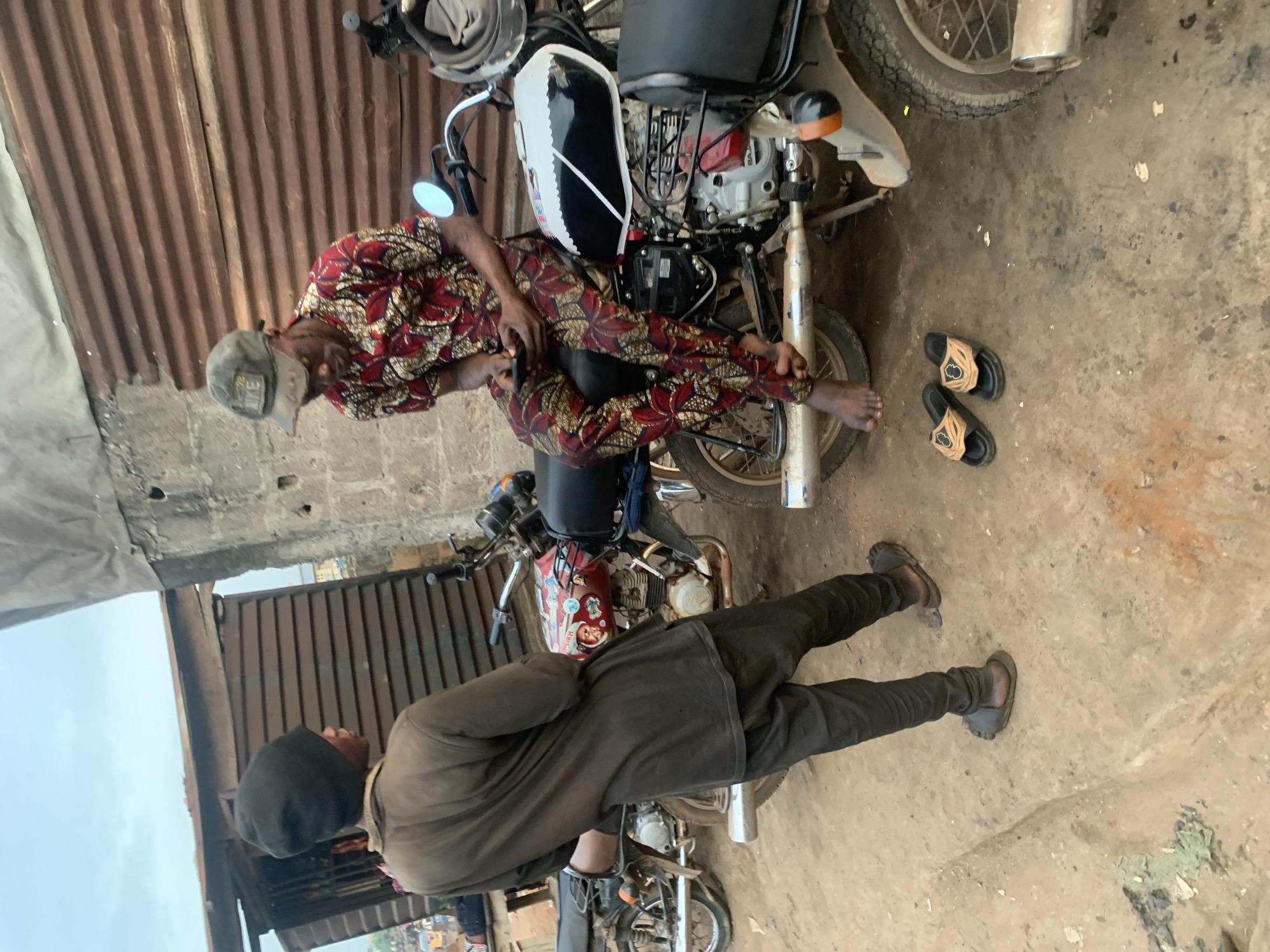 Two men are in an outdoor setting, likely a dusty roadside in Shagamu, Nigeria. The man on the left, wearing a dark beanie, a long-sleeved brown shirt, and dark pants, stands facing away from the camera, looking towards a row of brown corrugated metal structures that appear to be shops or sheds. He is wearing dark sandals.
The man on the right is seated on a motorcycle, dressed in a vibrant red and gold patterned shirt and matching patterned pants. He is wearing a grey baseball cap and is holding a mobile phone, looking down at it. His bare feet are visible. Another motorcycle is parked behind him, and a third is partially visible to the left. Flip-flops are placed on the ground near the motorcycle the man is sitting on. The ground is a mix of dirt and sand, with some patches of green and orange material scattered on it. The overall atmosphere suggests a casual, everyday scene in a bustling urban environment.
