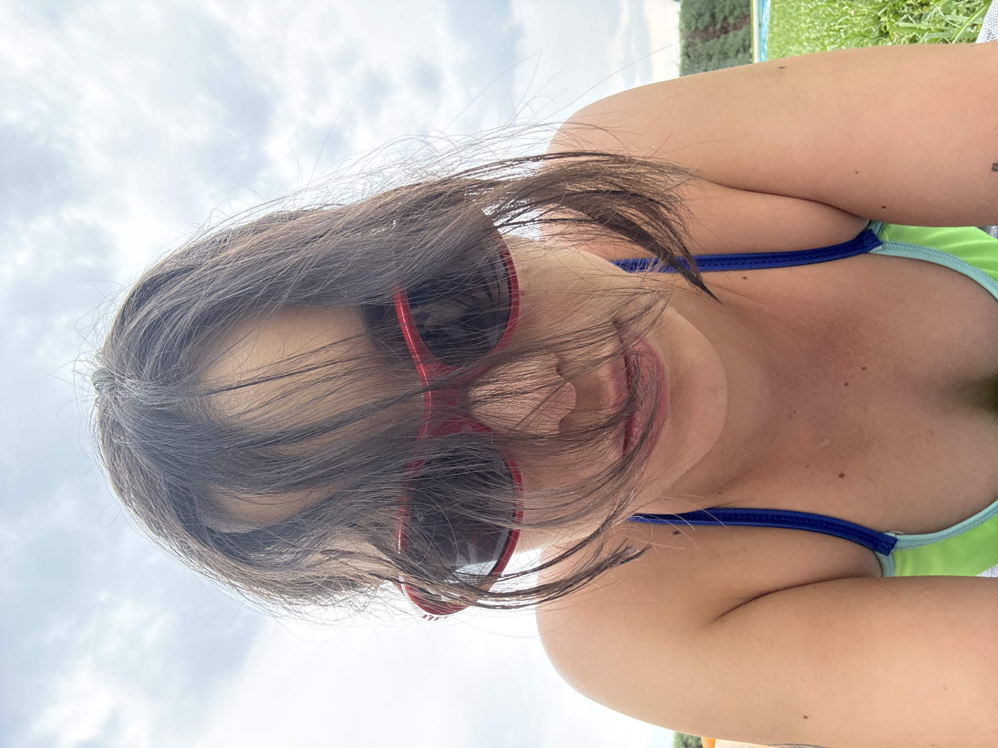 A close-up, low-angle, selfie-style photograph depicts a young woman wearing red heart-shaped sunglasses and a swimsuit. Her dark brown hair partially obscures her face. The woman appears to be outdoors, possibly near a swimming pool, as suggested by the blurred background which includes grass and a body of water. The sky is overcast with clouds. The specific location in El Escorial, Spain is not discernible from the image itself.