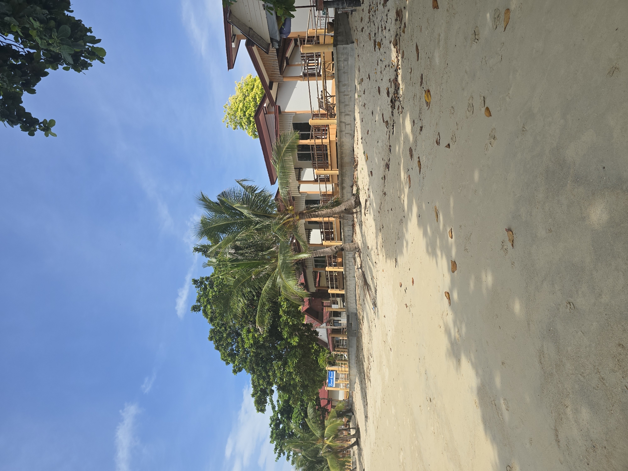 The image depicts a tropical beach scene at Lamai Resort in Baan Lamai, Thailand. The foreground is dominated by a sandy beach with scattered fallen leaves and footprints. Behind the beach, a row of bungalows with orange-tiled roofs and white walls are visible, each featuring a wooden porch. Palm trees and lush green foliage line the area between the bungalows and the beach. The sky is a clear blue with a few wispy clouds. A sign for "Lamai Resort" is present in the middle ground. The scene suggests a tranquil resort setting on a sunny day.