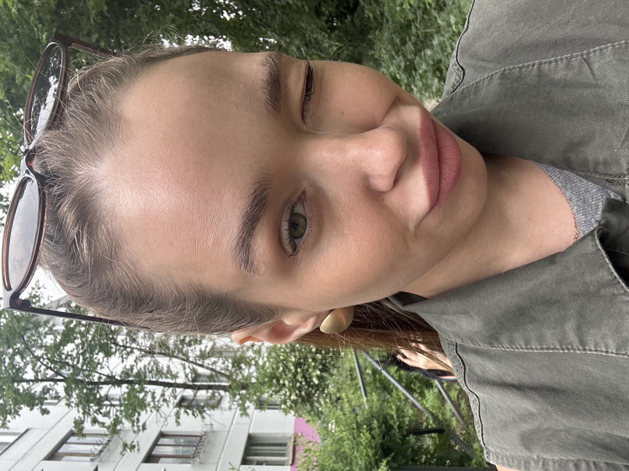 A young woman with light brown hair and a tan complexion is taking a selfie outdoors. She is wearing a green collared shirt over a grey t-shirt and large gold earrings. Her sunglasses are resting on top of her head. She is winking and making a puckered face. The background shows a building, trees, and a railing. The city of Chaikovsky, Russia, is known for its hydroelectric power station and surrounding natural beauty, suggesting the woman might be near a residential area or park within the city.