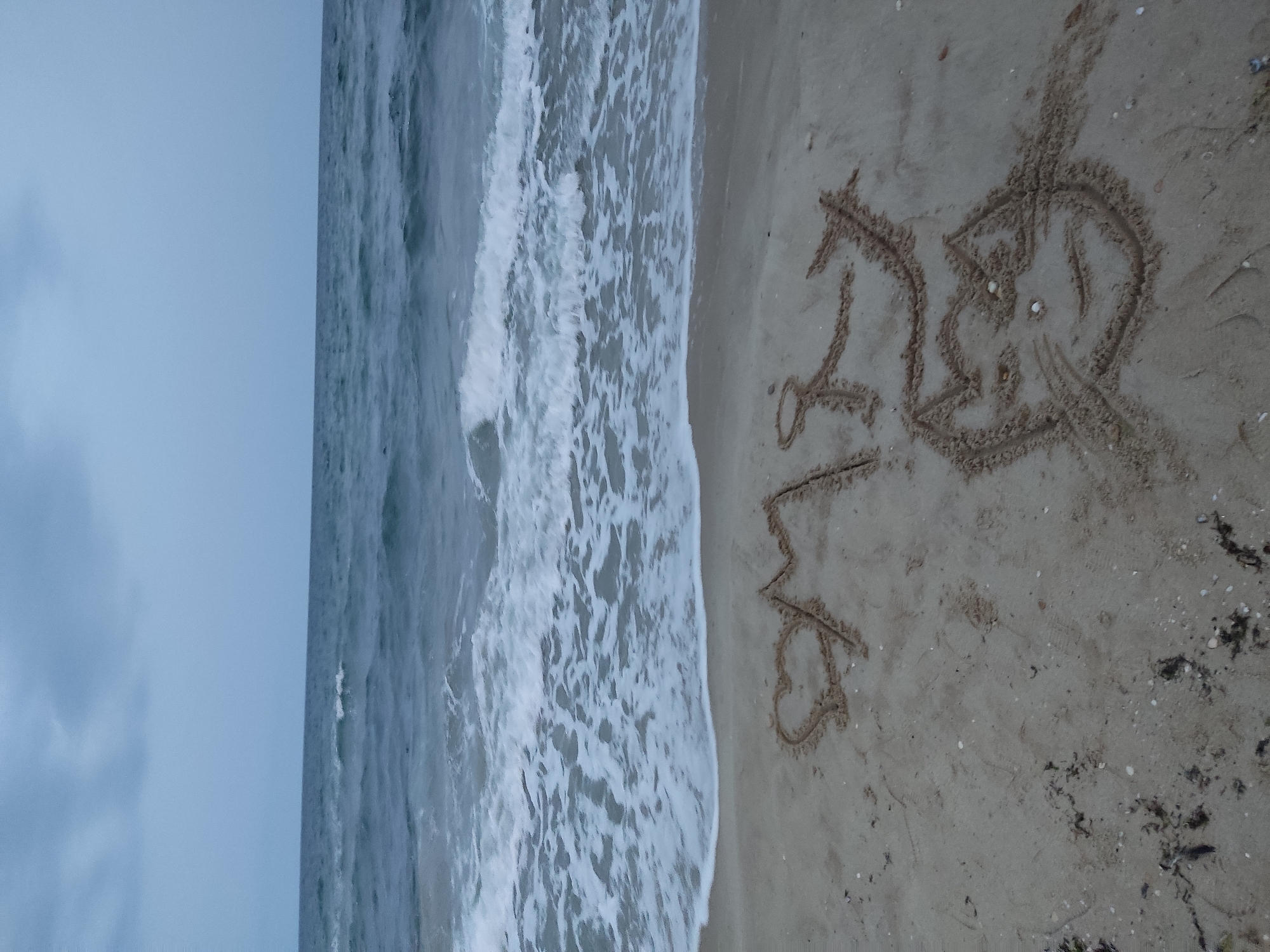 The image depicts a sandy beach meeting a body of water, likely the ocean. The word "DUB" is written in the sand, accompanied by a heart symbol. The water appears to be rough with visible waves. The sky is overcast. The location is an unidentified beach within the city of Unknown, Unknown. There are no people visible. Additional sand details, such as scattered shells or debris, are visible near the writing.