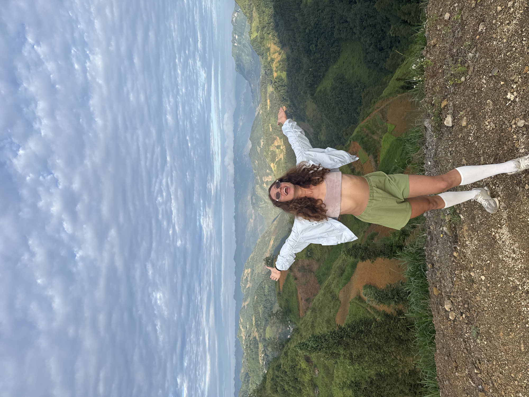 The image shows a person standing on a rocky precipice, appearing to be the edge of a mountain or elevated terrain, possibly in the Lũng Cú Commune, Vietnam. The individual, wearing a light-colored jacket, a light pink top, green shorts, and knee-high white socks, has their arms outstretched with thumbs pointing up. They are also wearing sunglasses, revealing what appears to be a smile or expression of excitement. The background features a scenic landscape of mountains, valleys, and terraced rice fields under a cloudy sky. The terrain consists of cultivated land and lush vegetation.