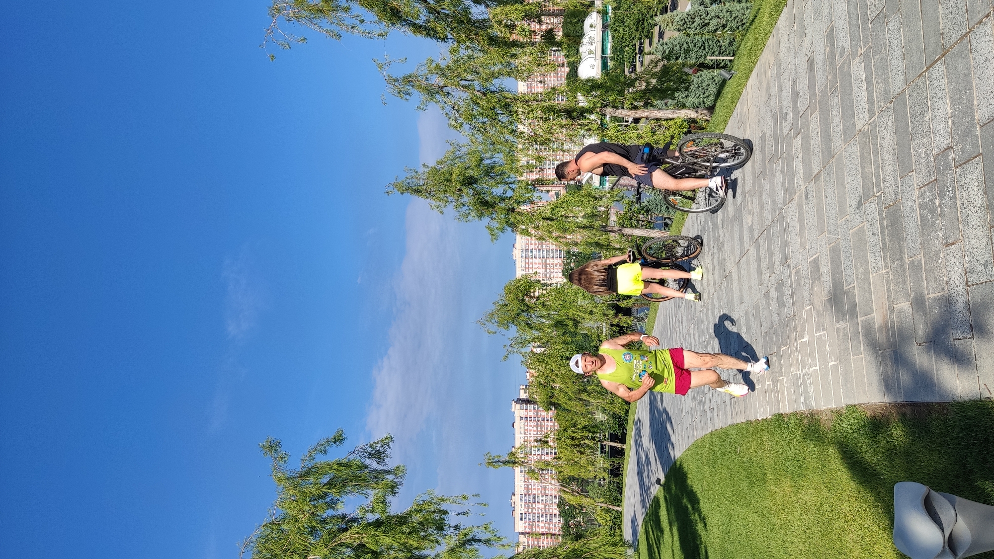 In Krasnodar, Russia, a man in a green tank top and red shorts runs along a paved park path under a clear blue sky. He wears a white cap and athletic shoes. To his right, a woman in yellow shorts and a black tank top rides a bicycle. Further to the right, a man in a black tank top and blue shorts also rides a bicycle. The path curves through a green park with manicured trees and grass. In the background, several multi-story apartment buildings are visible, suggesting an urban setting. The lighting indicates it is daytime, likely morning or afternoon.