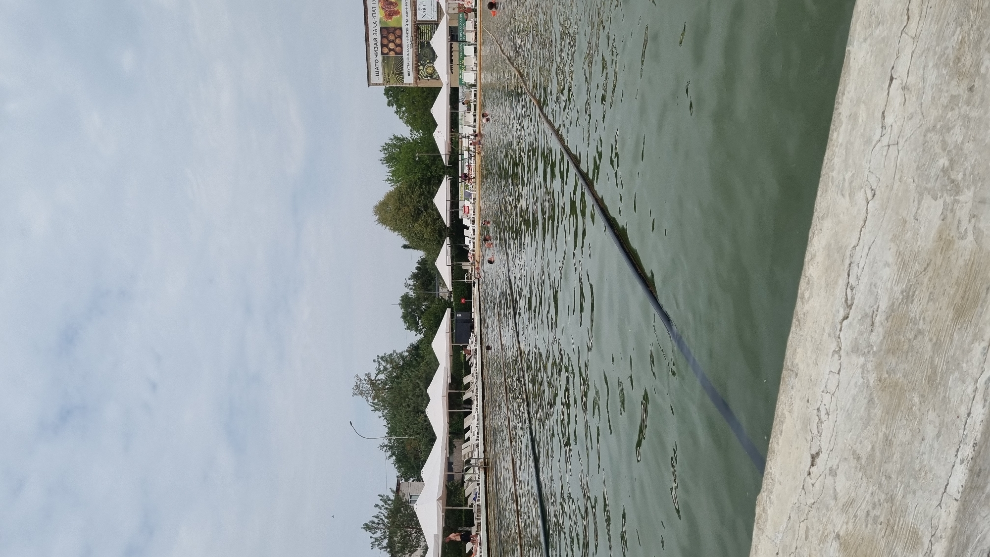 The image depicts an outdoor swimming pool facility in Berehove, Ukraine. In the foreground, the edge of the pool is visible with a concrete border. The water in the pool is a murky green color, and a black rope floats on the surface, dividing lanes. Several people are seen swimming or wading in the pool. Along the far edge of the pool, a row of white umbrellas provides shade for lounge chairs. Behind the lounge chairs, there are buildings and trees. A large billboard with text and images is visible on one of the buildings, suggesting this is a commercial establishment. The sky is overcast with scattered clouds. The overall impression is of a public recreational area on a cloudy day.