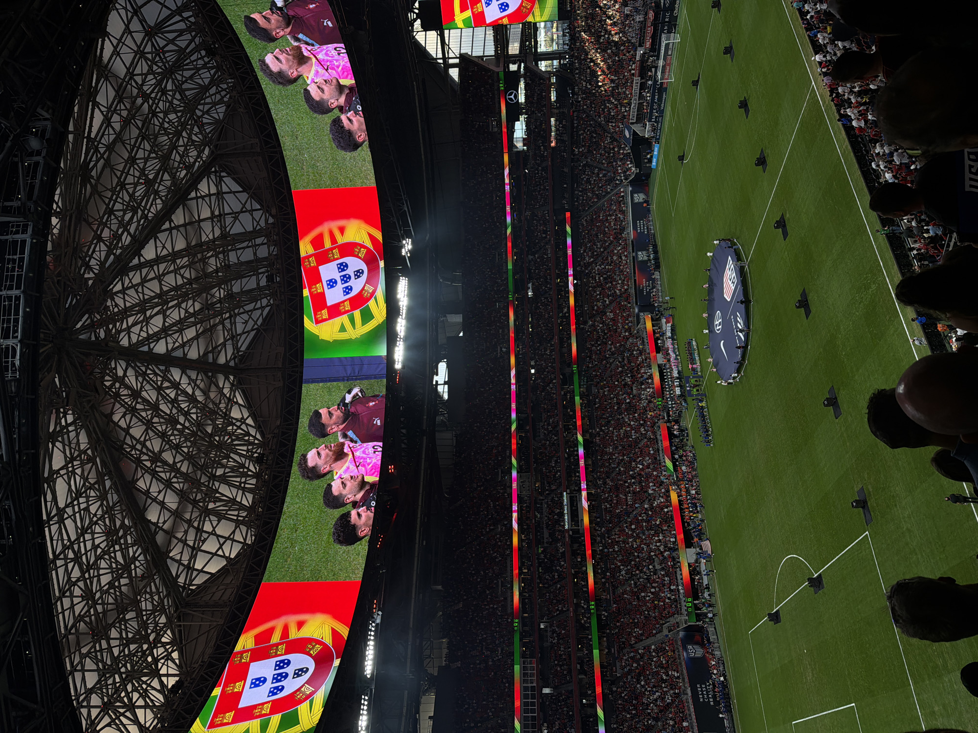 The image captures a view inside Mercedes-Benz Stadium in Atlanta, United States, during what appears to be a soccer (football) match or pre-match ceremony. The stadium is filled with spectators, visible in the stands surrounding the field. A large, circular video screen is suspended above the field, displaying images including a group of players and the flag of Portugal. The soccer field is green and marked with white lines. In the center of the field, there's a large round cover with an emblem (likely related to one of the teams). In the foreground, the heads of some spectators are visible. The architecture of the stadium roof is visible, constructed of a complex web of steel beams.