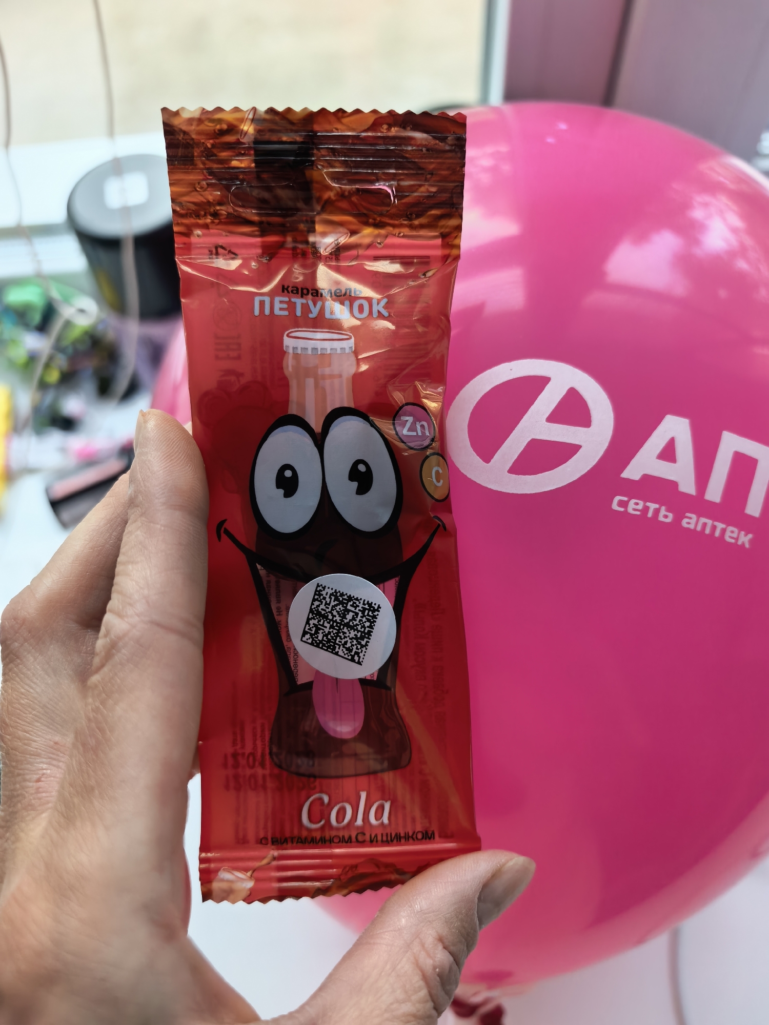 A person is holding a red caramel lollipop packet with a cartoon cola bottle design, including large eyes and a QR code sticker. The package reads "Cola" and "С ВИТАМИНОМ С И ЦИНКОМ," which translates to "With Vitamin C and Zinc." In the background, a pink balloon with a logo is visible, featuring a circle with an 'A' inside and the text "ΑΠ сеть аптек" (AP Pharmacy Network). A partially visible window and other objects are blurred in the background. The text on the packet also includes the word "карамель" (caramel) and "ПЕТУНОК" (Petushok). A sticker with a QR code is attached to the packaging, covering part of the design. In the barcode next to Cola is written "45045058 TDIST". The scene is likely in Sterlitamak, Russia, considering the text is in Russian and the presence of a pharmacy network.