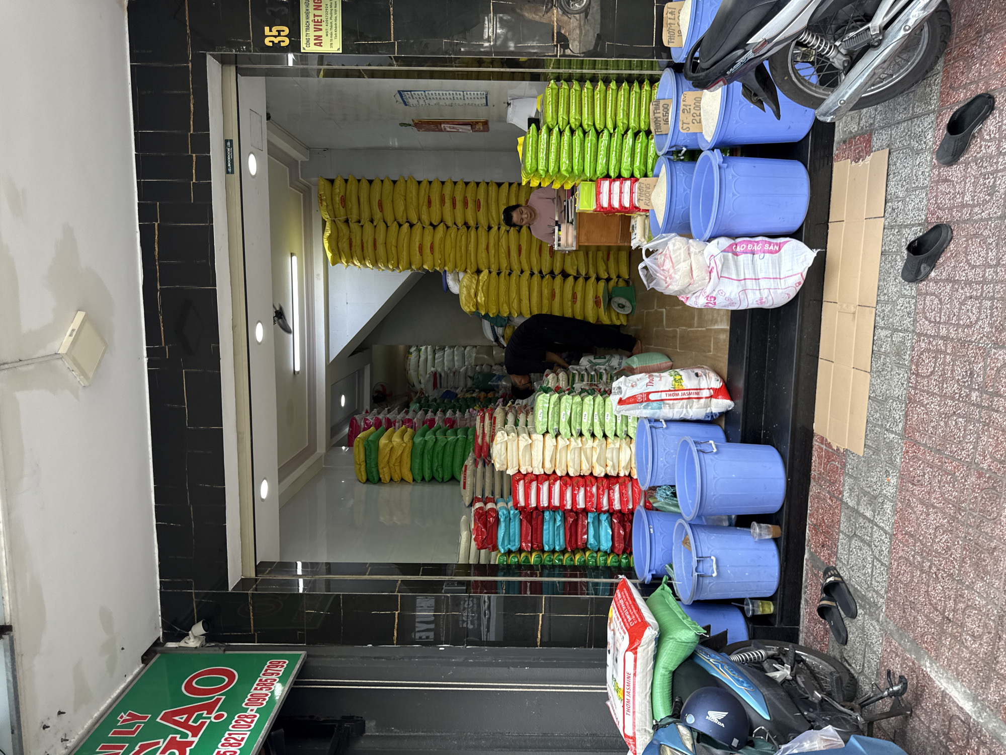 The image depicts the storefront of a rice shop in Nha Trang, Vietnam. Large stacks of bagged rice, in various colors including yellow, green, red, and white, are prominently displayed both inside and outside the store. A motorcycle is parked on the left side of the frame, partially obstructing the view of the entrance. Several large blue plastic buckets are lined up in front of the store, some containing rice. Two pairs of black sandals are placed on the tiled sidewalk in front of the entrance. Inside the store, a woman is seated at a desk, and another person, dressed in black, is visible in the background, interacting with the rice bags. A sign on the left side of the building is green with red lettering. Another sign on the right displays the number "35" and Vietnamese text. The environment appears to be a commercial street.