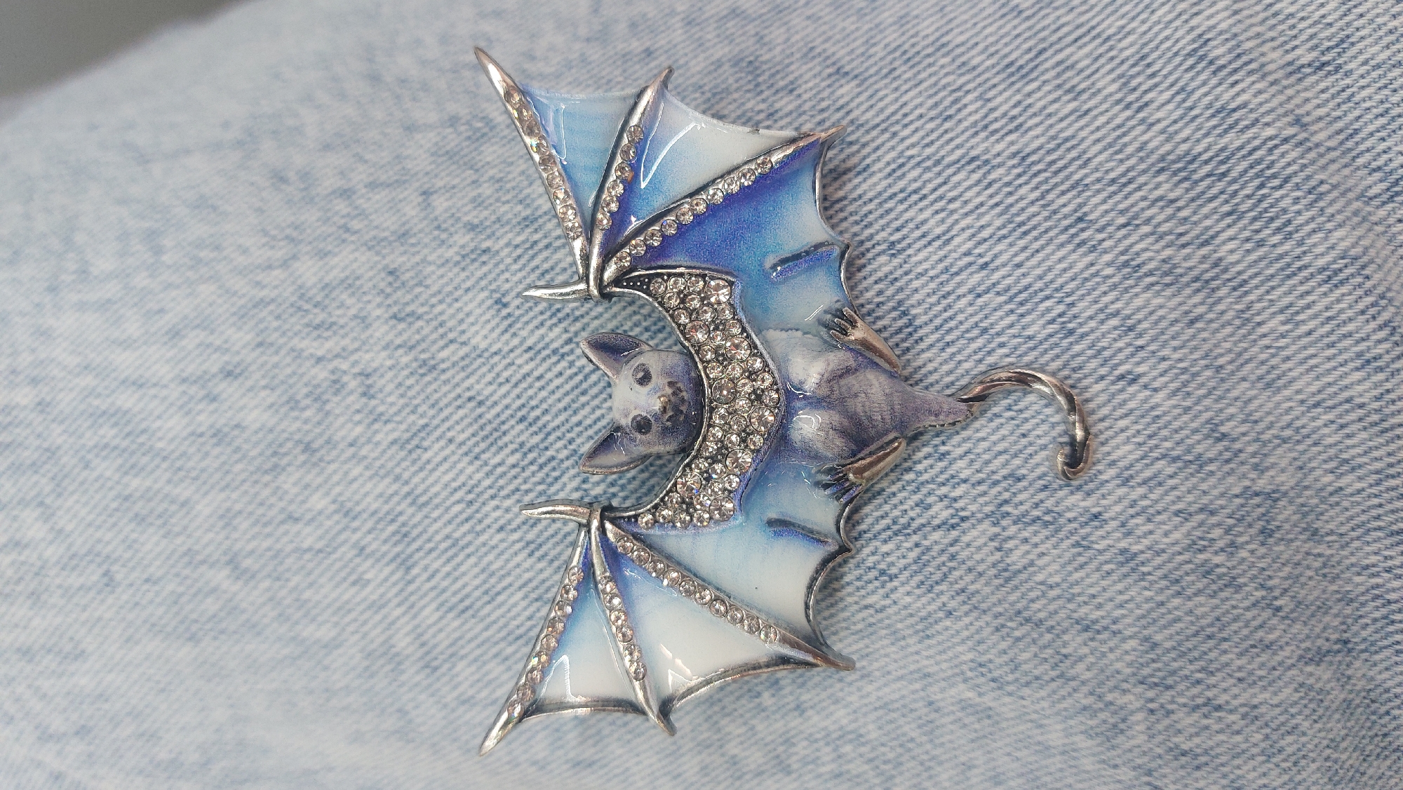 The image shows a decorative bat-shaped brooch, likely made of metal and enamel. The brooch is embellished with small, clear crystals along the wing edges and body. The enamel coloration transitions from white to light blue to a darker blue. The bat has a curved tail. The brooch is lying on a light blue denim fabric with a distinct weave pattern. There are no indications in the image that reveal a connection to Novosibirsk, Russia.