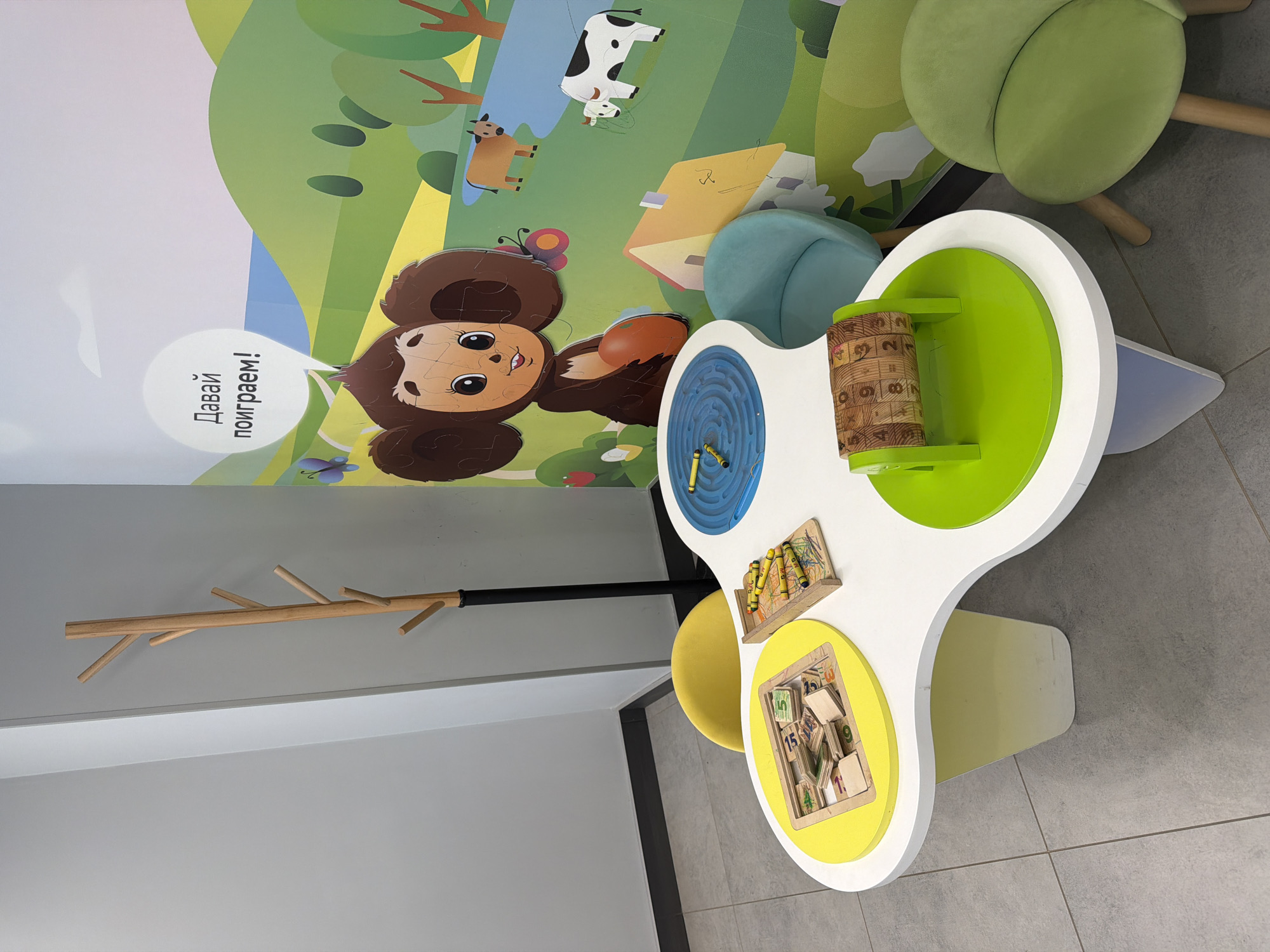 The image shows a children's play area, possibly in a public space like a clinic or shopping mall, located in Chaikovsky, Russia. A wall mural features cartoon farm animals and a large puzzle image of a cartoon character with a speech bubble saying "Давай поиграем!" (Let's play!). A white, clover-shaped table with colorful insets (blue, green, and yellow) has various games and activities, including a maze, a cylinder with numbers, a crayon holder with crayons, and a container with wooden blocks with letters and numbers. A green chair sits near the table, and a wooden coat rack is mounted on the wall nearby. The floor is tiled in gray.