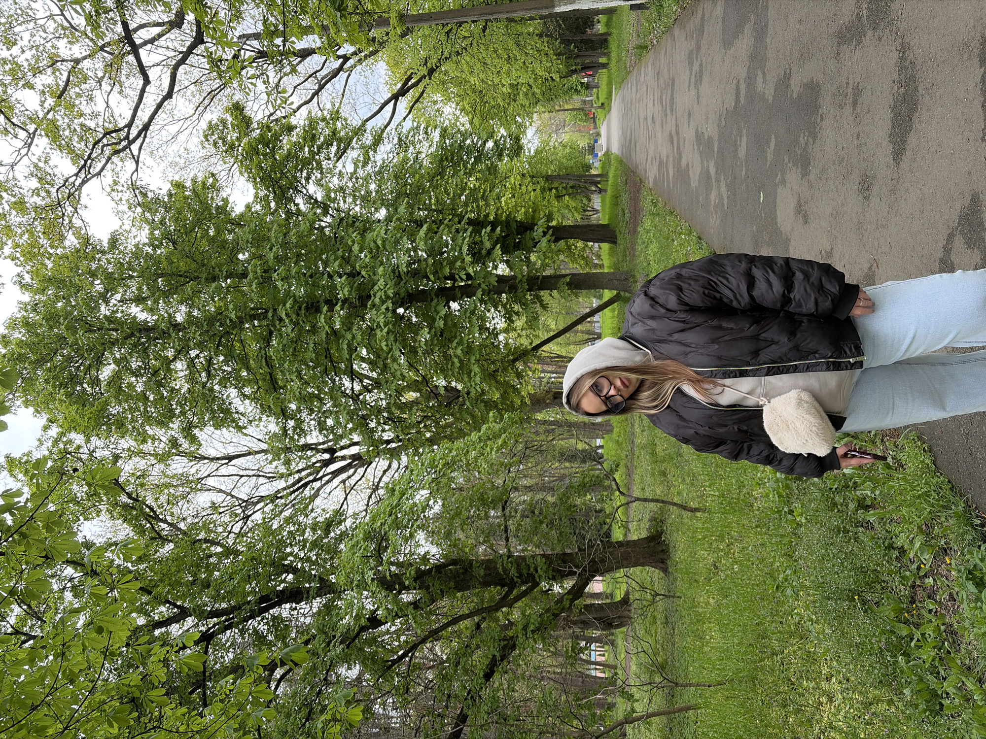 A young woman is captured in a park setting, likely in Krasnodar, Russia, during what appears to be a bright spring or early summer day. She stands on or near a paved pathway, amidst a dense canopy of lush green trees, whose new leaves signify the season. The ground is covered in vibrant green grass and some smaller plants.
The woman is the central subject, looking towards the camera with a relaxed demeanor. She wears large, dark-framed glasses, and her long, light-colored hair with hints of pink highlights frames her face. Her attire includes a light grey hooded top, a black puffer jacket, and light blue jeans. A small, fluffy, cream-colored handbag hangs from her shoulder. Her left hand holds a dark-colored smartphone, resting by her side.
The natural light suggests it is daytime, with the sky appearing bright and clear or lightly overcast through the tree branches. The overall atmosphere is serene and natural, characteristic of a leisurely stroll or an outdoor portrait in a park. There is no visible text in the image.