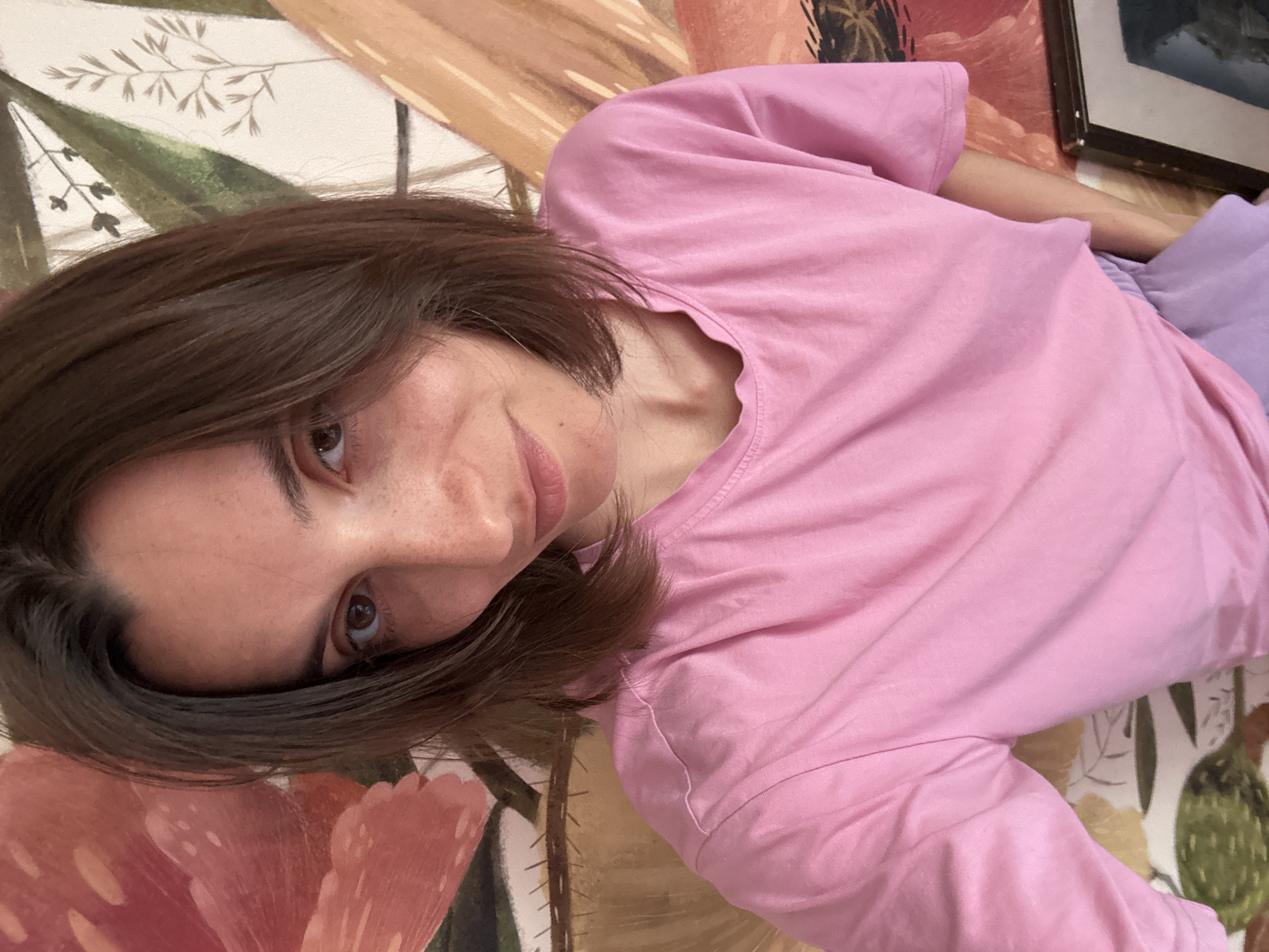A woman with brown hair is lying on a floral-patterned surface. She is wearing a pink t-shirt and purple shorts. The surface appears to be a bedspread or blanket. A framed picture is visible in the background. There is no indication of the location being Moscow, Russia.