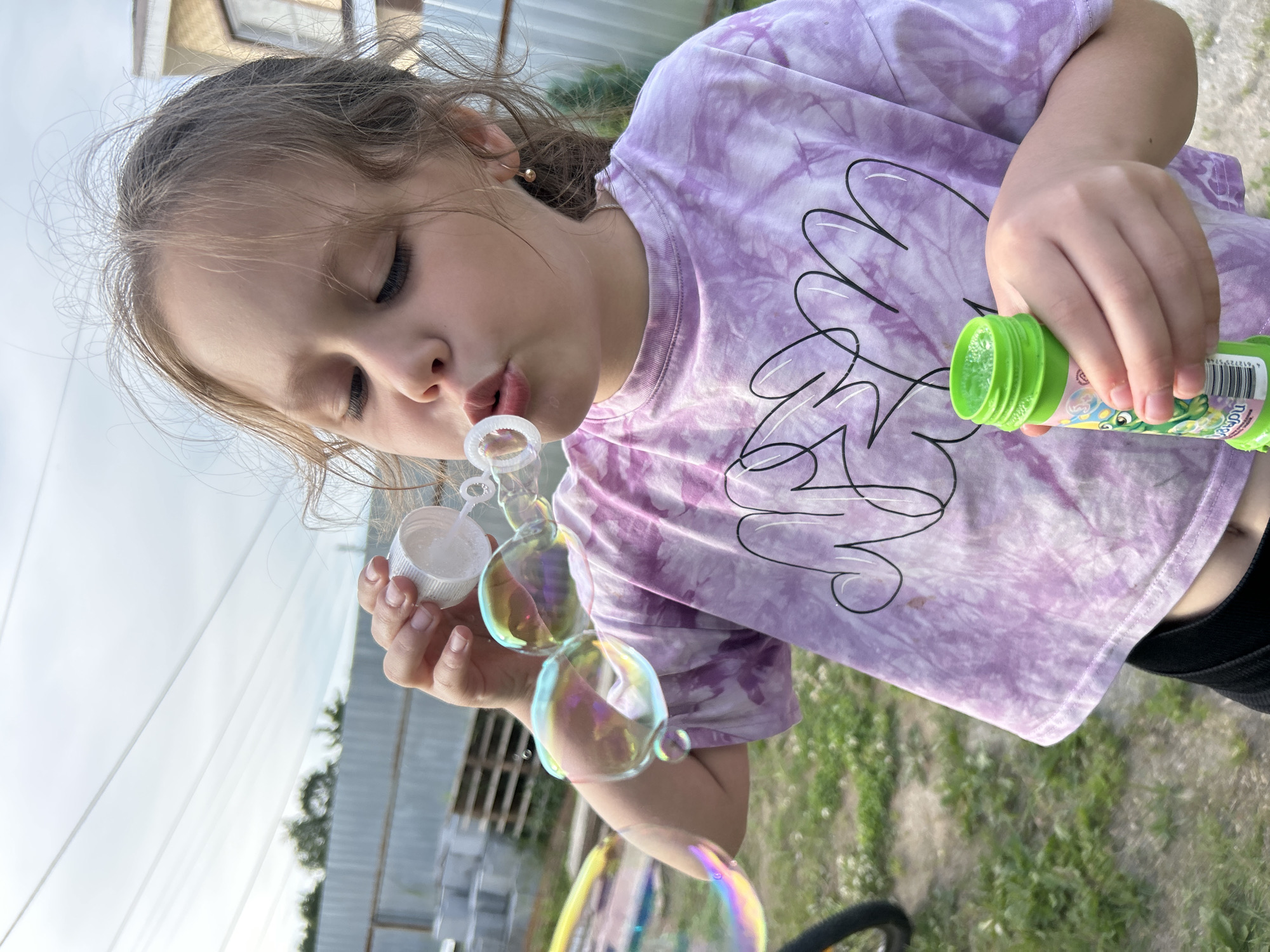 A young girl is blowing bubbles outdoors. She is wearing a purple tie-dye t-shirt with a black outline drawing of a flower on it. She holds a green bottle of bubble solution in her right hand and is blowing through a bubble wand held in her left hand. Several iridescent bubbles are floating in the air around her. Her eyes are closed and her lips are pursed as she blows. The setting appears to be a backyard with grass and some fencing in the background. The lighting suggests it is daytime, possibly late afternoon. The image is oriented vertically.