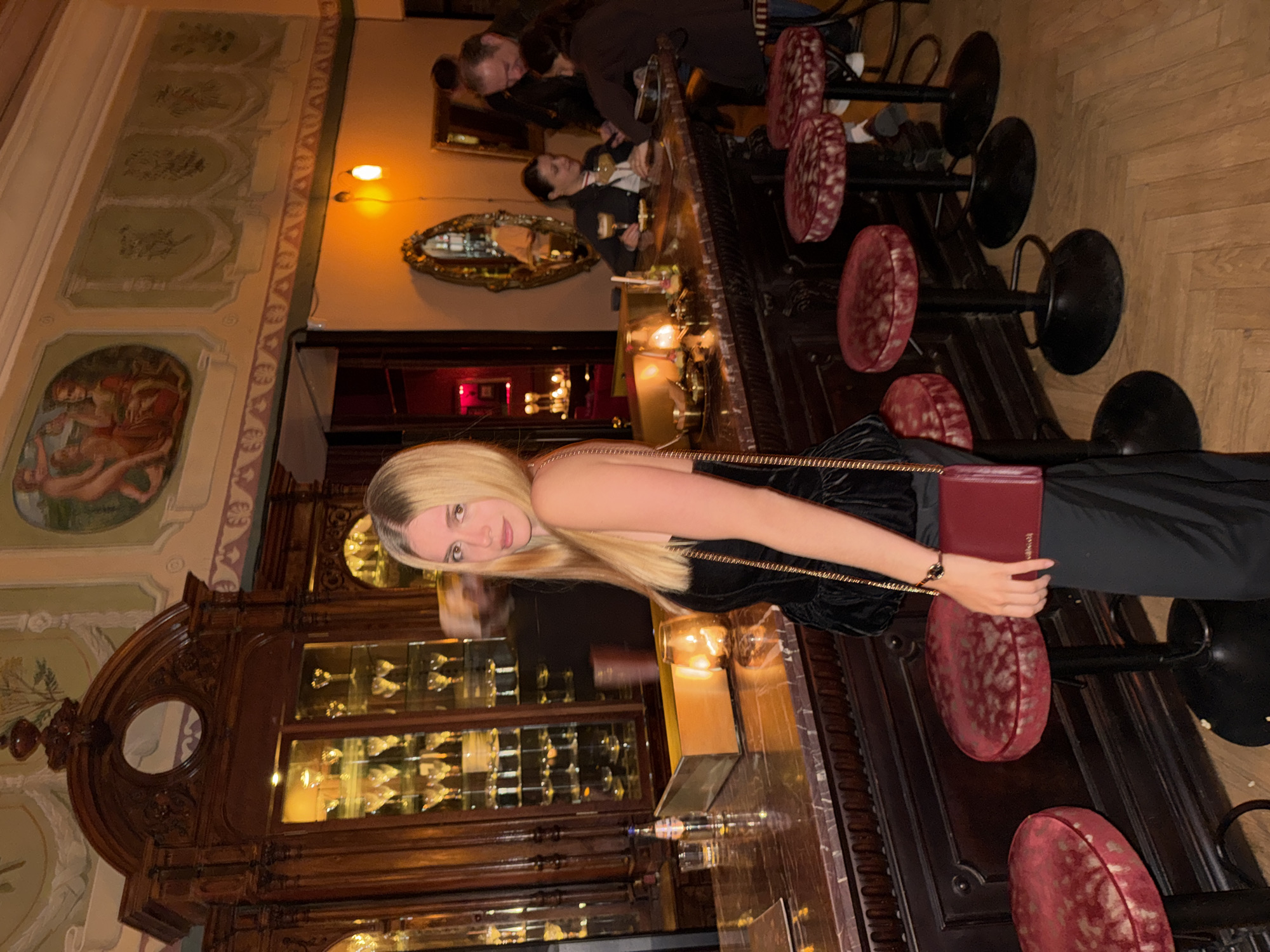 The image shows a blonde woman at a bar with red stools. She is wearing a black dress, holding a red clutch, and facing the camera. Behind her is a bar with a glass display case filled with bottles. In the background, there are other patrons at the bar and decorative elements on the walls. The painting on the wall and ornate architecture suggest a historic or upscale establishment. The location is most likely in Tbilisi, Georgia, given the context.