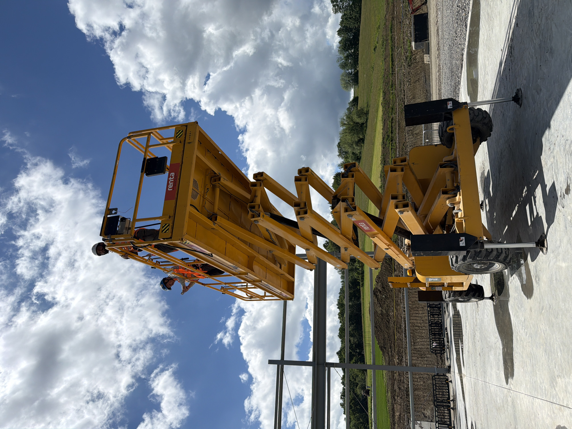 In Janowice, Poland, a construction scene unfolds under a partly cloudy sky. A bright yellow scissor lift, branded with "Renta," is raised, its platform extending towards unfinished steel building structures. Two construction workers, clad in orange safety vests and hard hats, occupy the platform. One worker, on the left, is looking down at a device in their hand, while the other, on the right, stands looking forward. The lift's machinery is visible, with its hydraulic arms extended. The ground is a mix of concrete and gravel, with puddles reflecting the sky. In the background, a grassy hill rises, dotted with trees. A tractor is faintly visible in the distance on a field. The overall impression is one of active construction work in a rural or semi-rural setting.