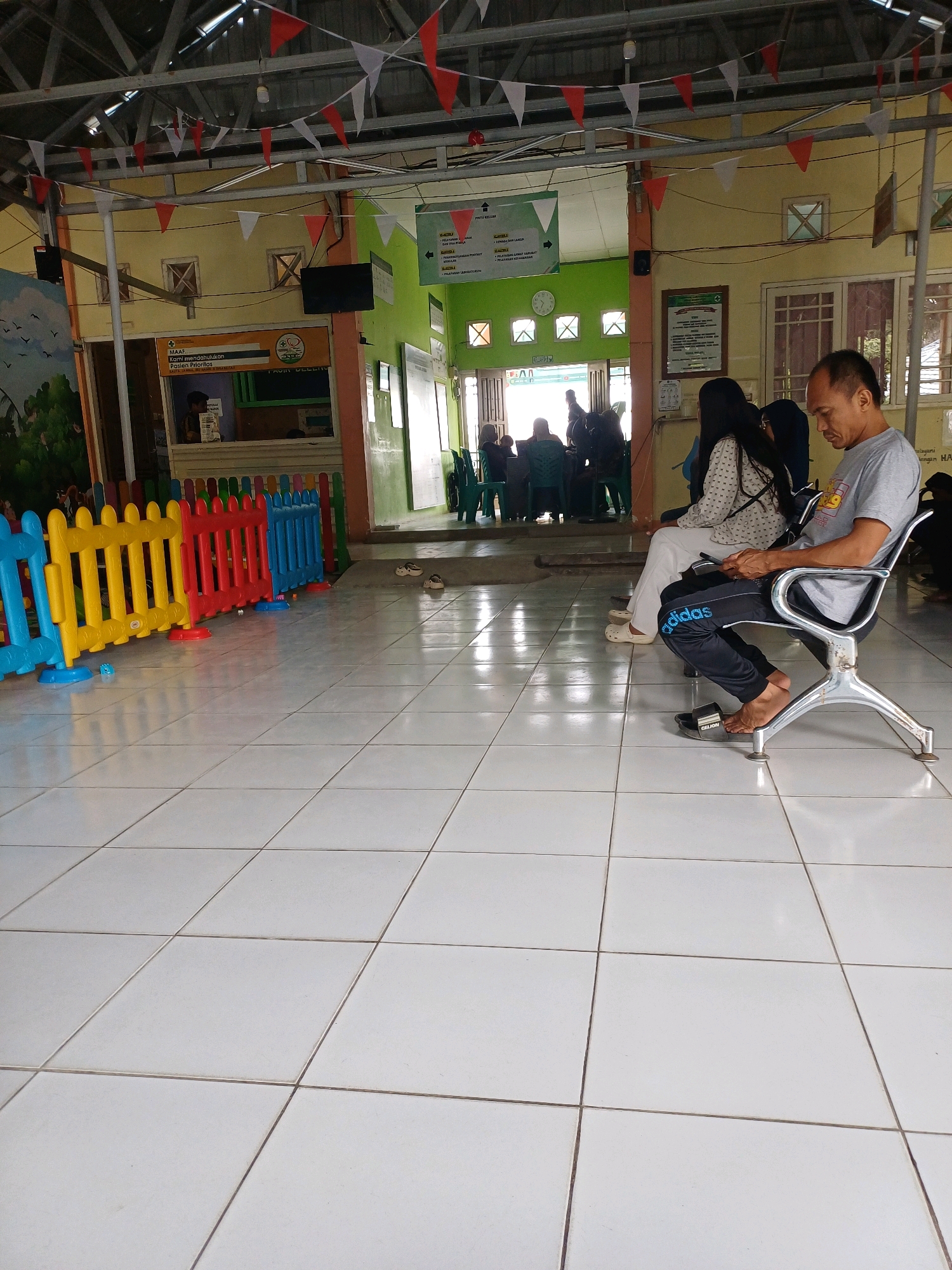 The image depicts the waiting area of a clinic or health center in Pasir Belengkong, Indonesia. In the foreground, the tiled floor is visible, with colorful plastic barriers in blue, yellow, red, and blue separating the waiting area from what appears to be an administrative or reception section. A man in a grey t-shirt and black pants with "adidas" written on them is seated in a metallic chair, looking down at his phone. Beside him, a woman in a patterned white top and white pants is also seated, looking away from the camera. Further into the background, several other individuals are seated on green chairs, and some are standing. The walls are painted yellow and green, and various signs and a clock are visible. Red and white triangular flags are strung across the ceiling. The overall environment suggests a public service facility.
