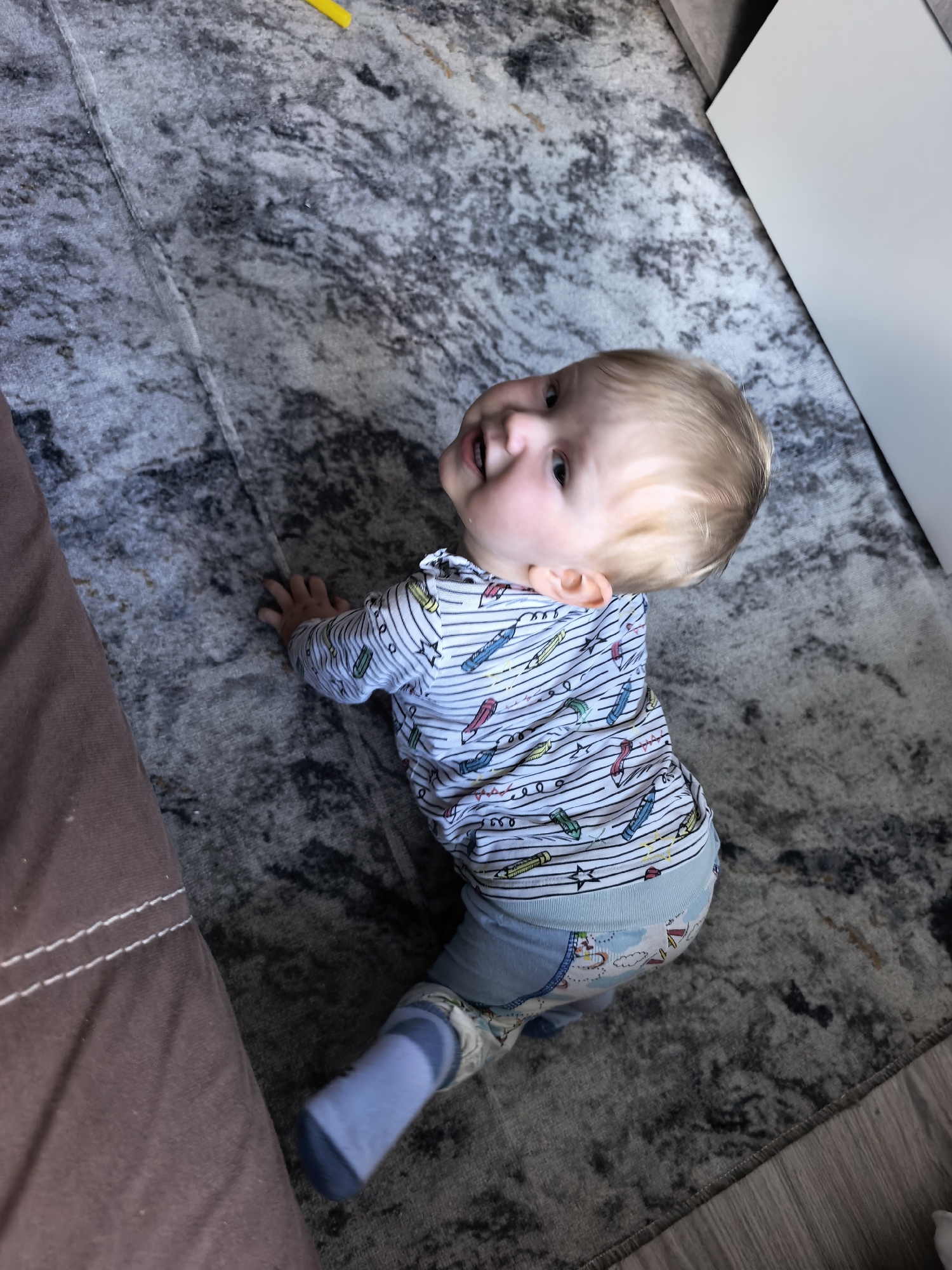 A toddler with blonde hair is crawling on a gray and white patterned rug. The toddler is wearing a blue and white striped shirt with crayon designs, blue pants with clouds, and a blue sock on their left foot. The rug is partially bordered by dark brown upholstery on the left side, and white furniture on the right. A yellow object is visible in the upper left corner of the image. The location in Sterlitamak, Russia cannot be determined from the image.