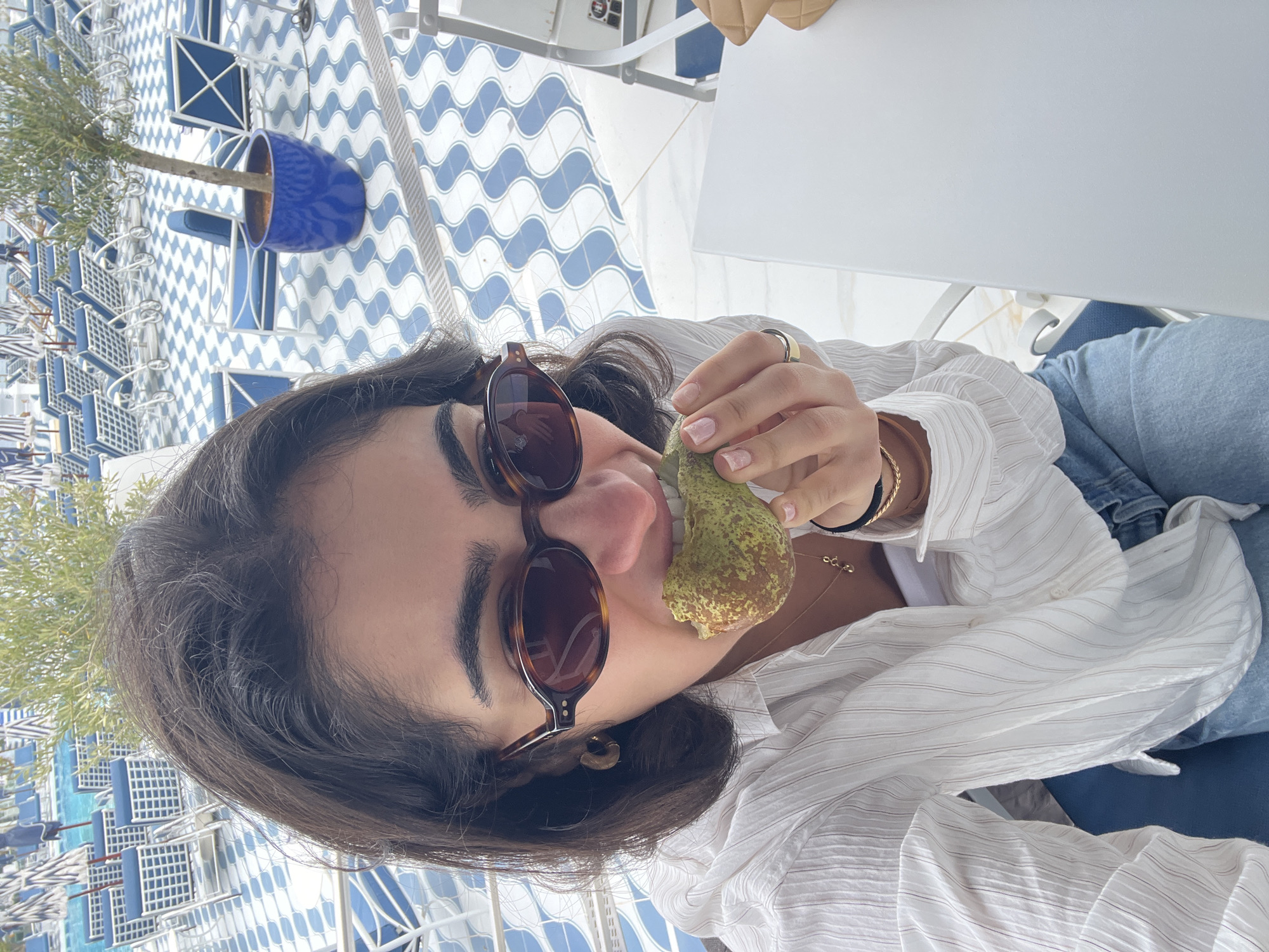 A woman wearing sunglasses and a white, striped shirt poses for a selfie. She is holding a pear up to her mouth. She is sitting at a white table on a blue and white tiled floor. A blue pot containing a small green plant is visible in the background. The location appears to be Santa Eulària des Riu, Spain.